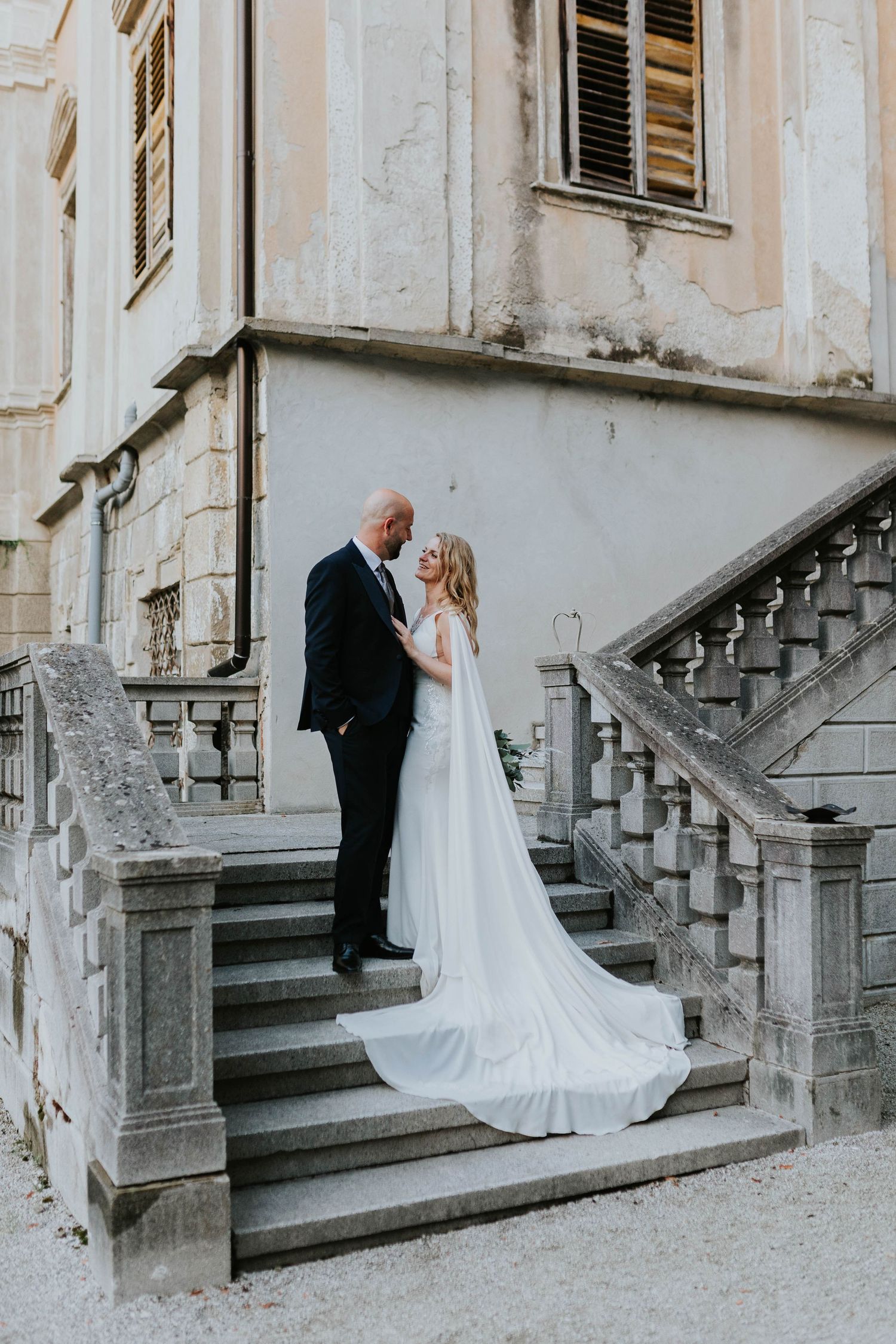 A bride and groom stand on stone steps of a historic building, sharing an intimate moment.