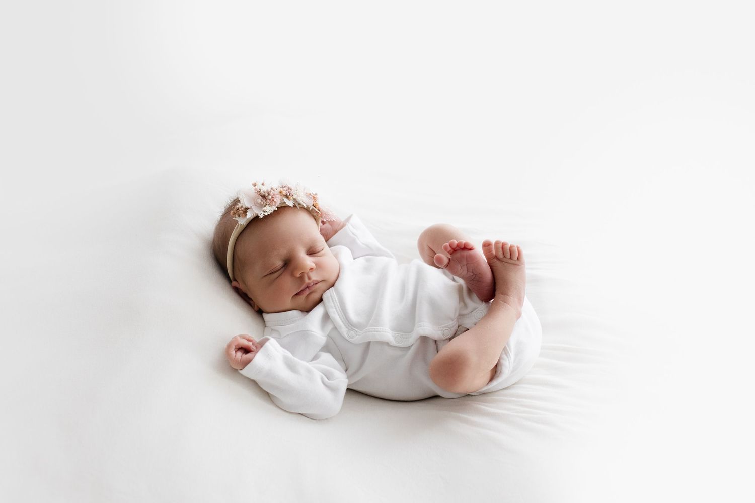 Sleeping newborn baby girl in white outfit with pink floral headband on white backdrop.