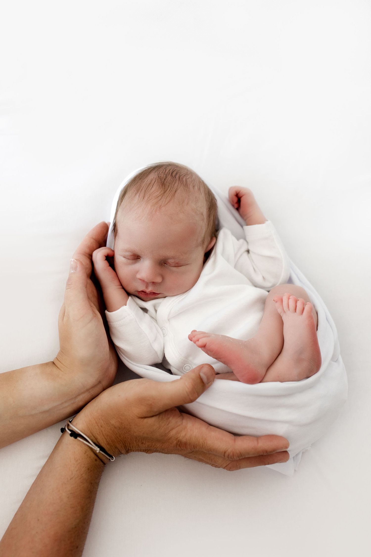 Sleeping newborn baby in white wrap cradled in parents loving hands on soft white background.