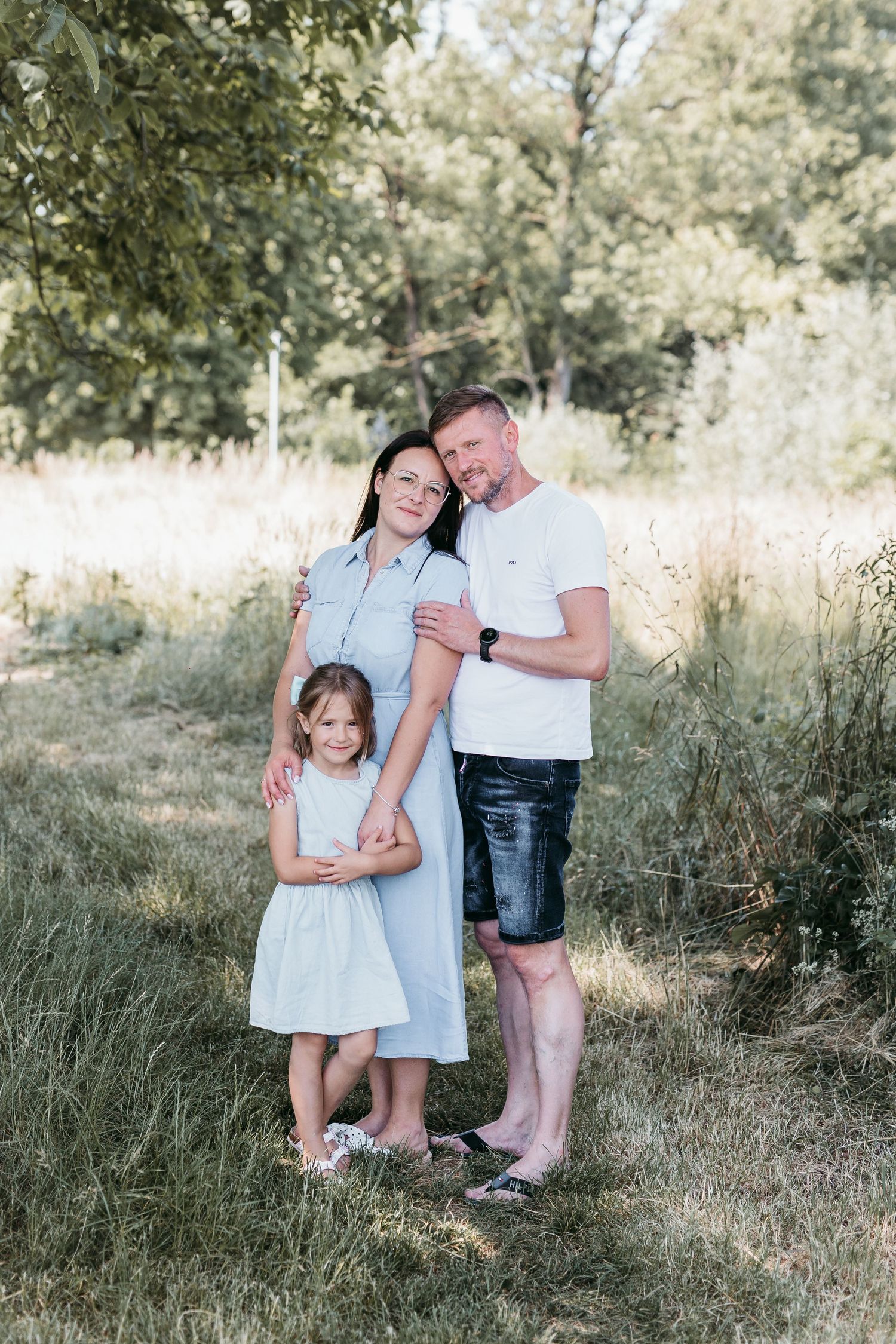 Family of three posing in a grassy field, parents embracing with young daughter standing in front wearing a white dress.