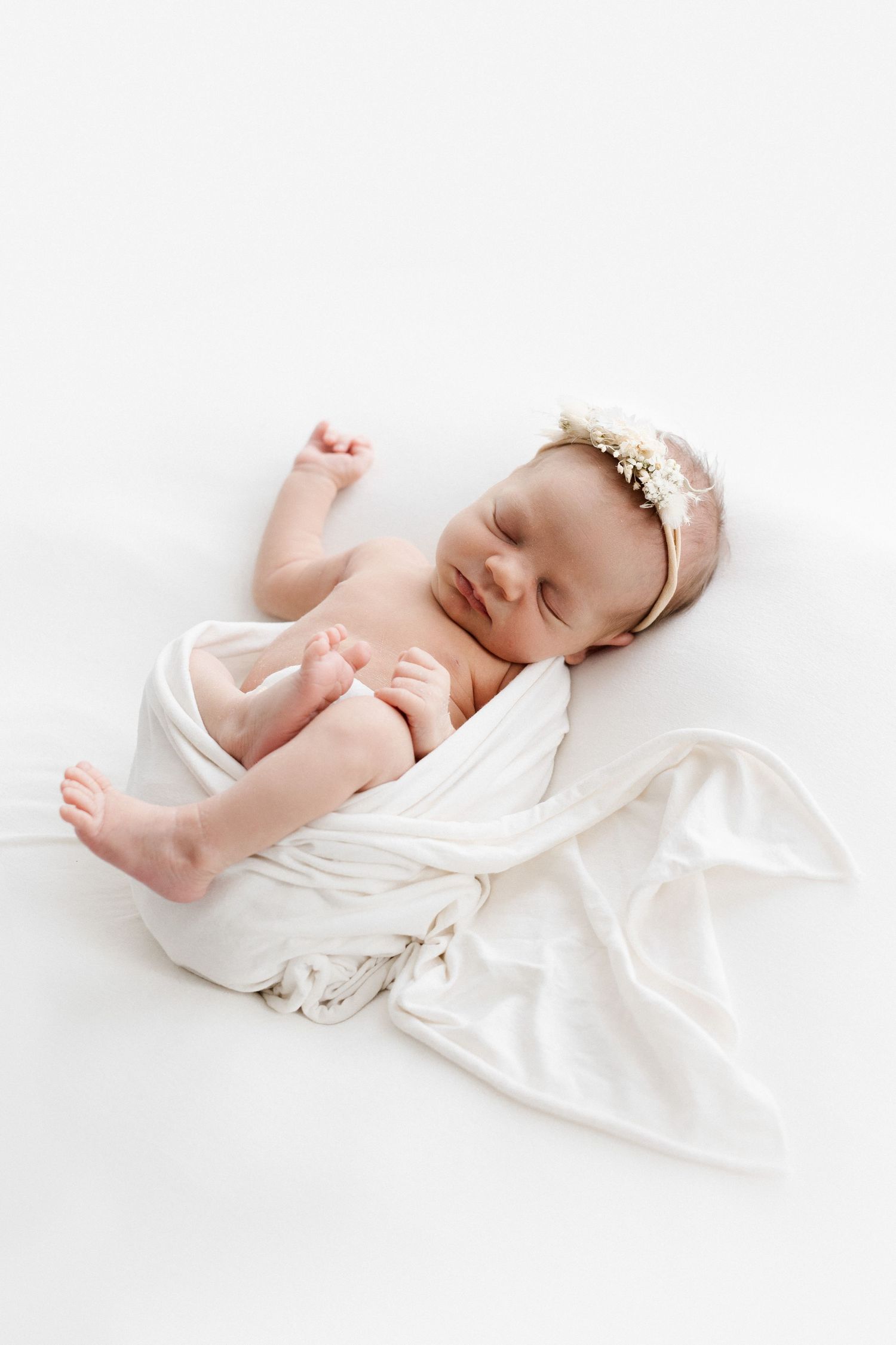 Sleeping newborn baby girl wrapped in white swaddle with gold floral headband on white background.