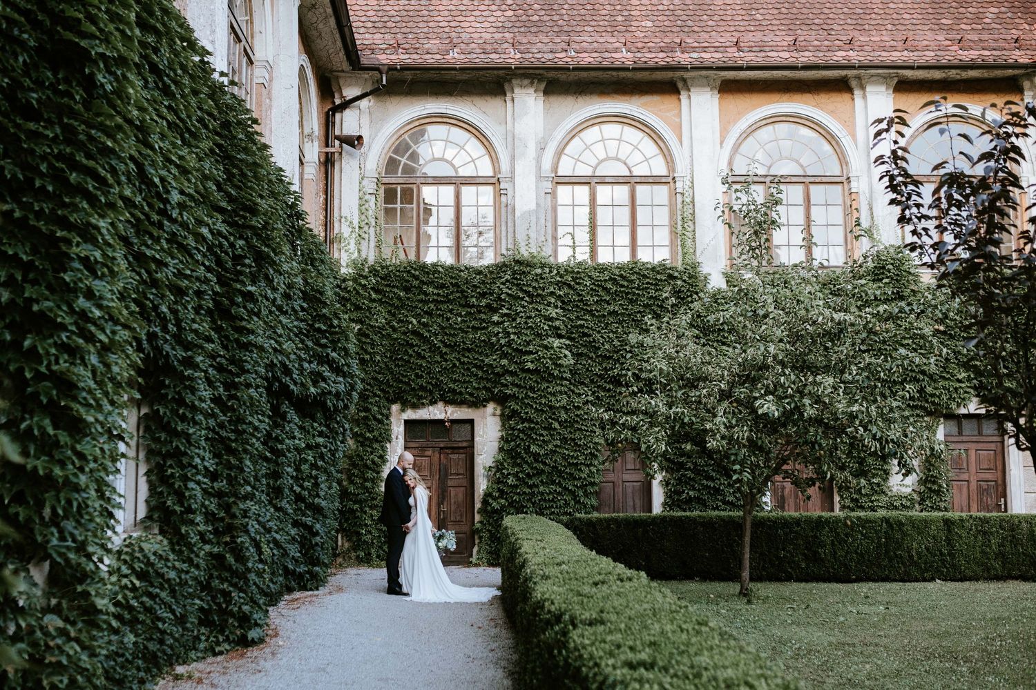 Bride and groom walking along a garden path beside an ivy-covered historic building with arched windows.