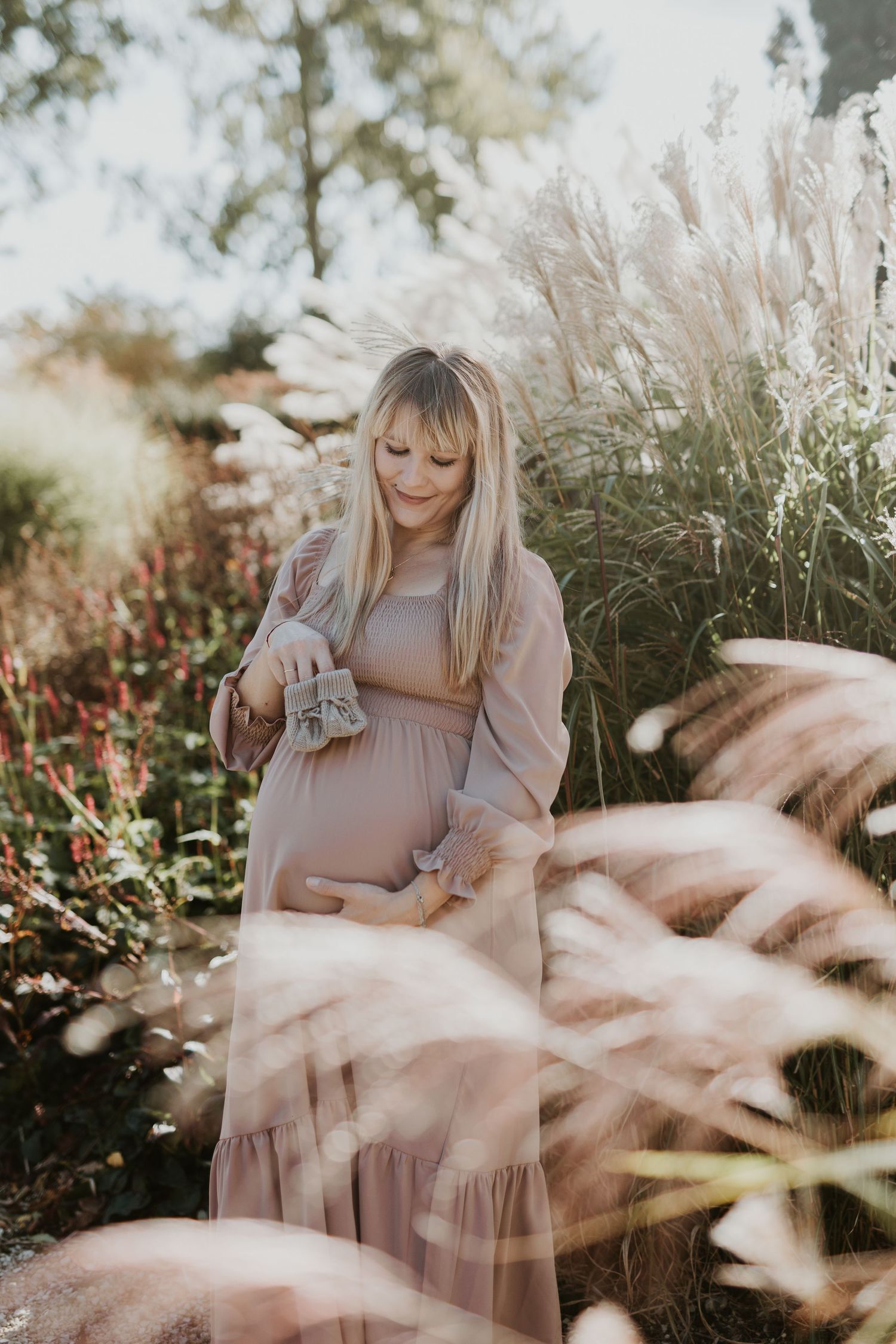 Pregnant blonde woman in mauve dress holding baby shoes in golden autumn garden.