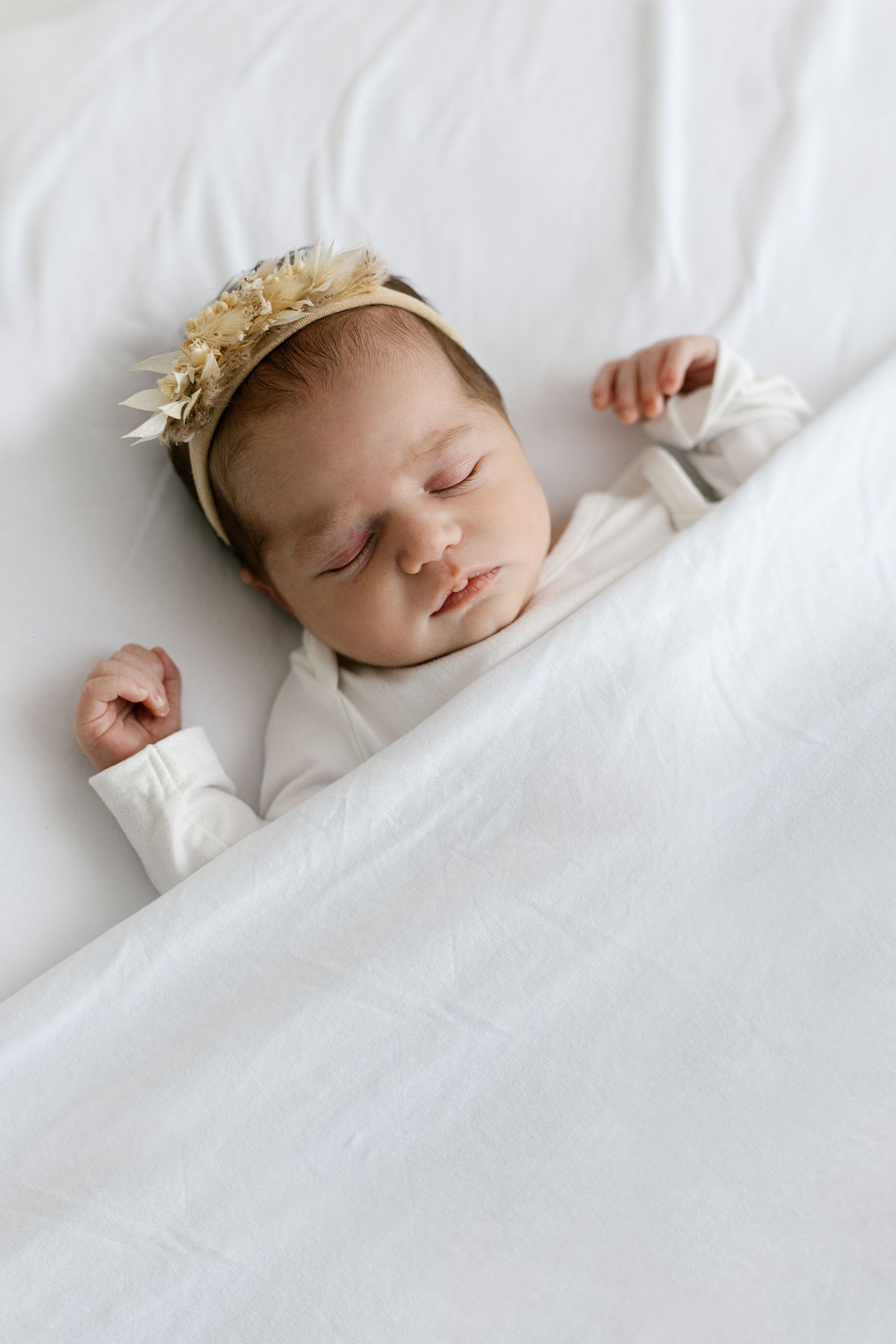 Peaceful newborn baby sleeping in white onesie with gold floral crown headband on white bedding.