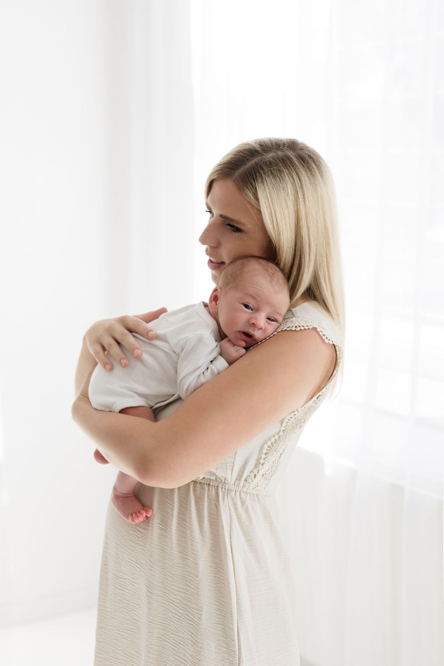 Blonde mother in white dress tenderly holds her newborn baby against her chest near a bright window.