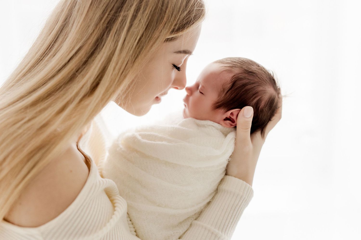 A blonde mother gently touches noses with her newborn baby wrapped in a white swaddle blanket.