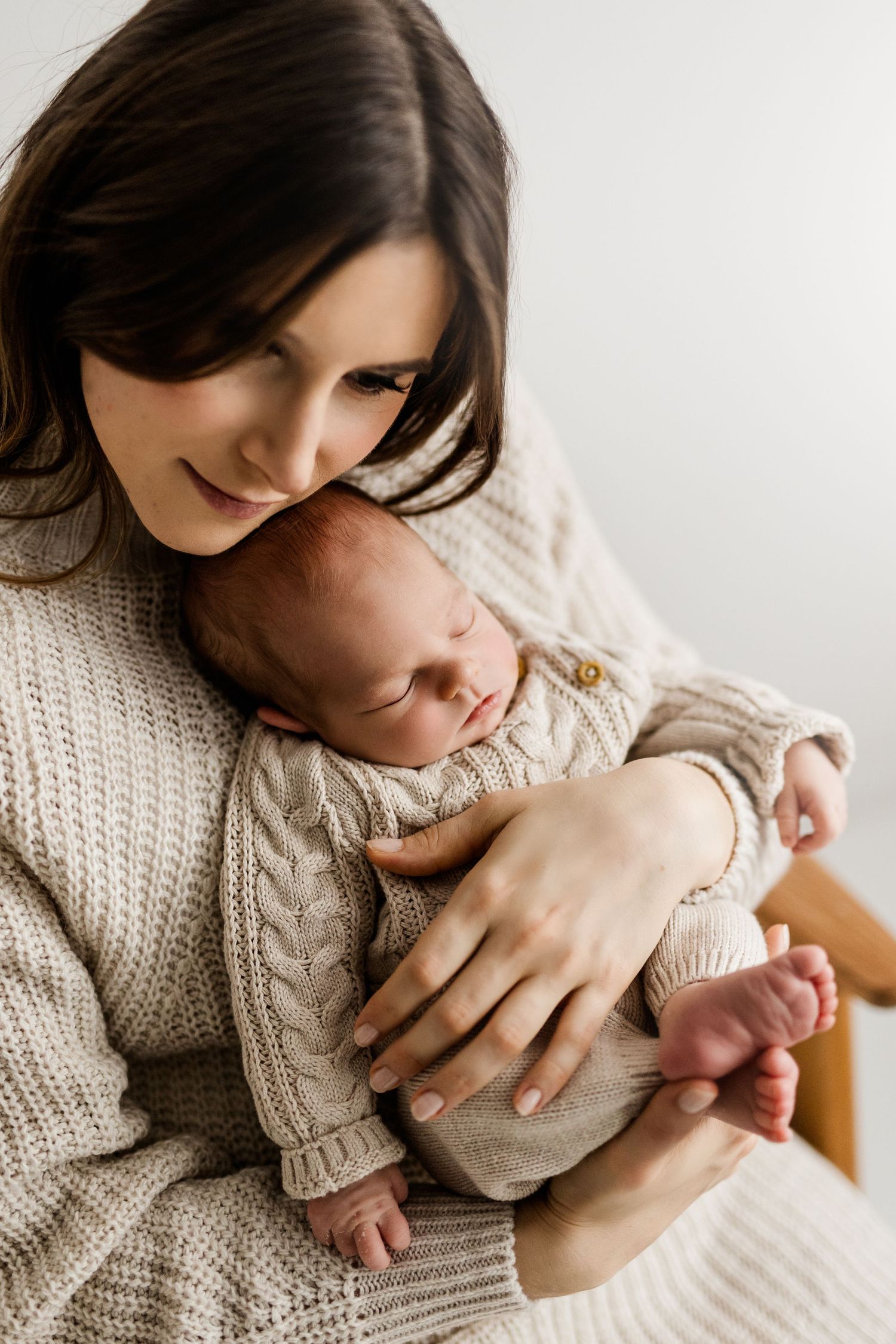 Dark-haired mother in cream knit sweater tenderly holding sleeping newborn baby.