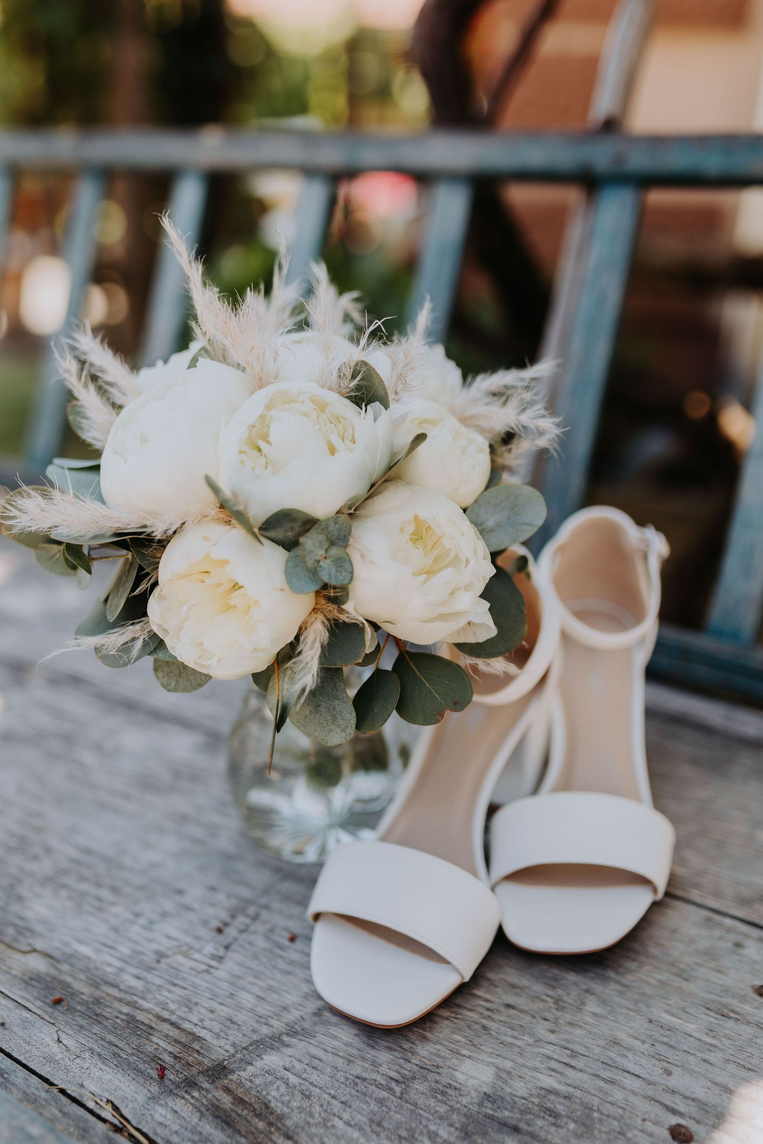 White bridal heels and peony bouquet with pampas grass displayed on a rustic wooden surface.
