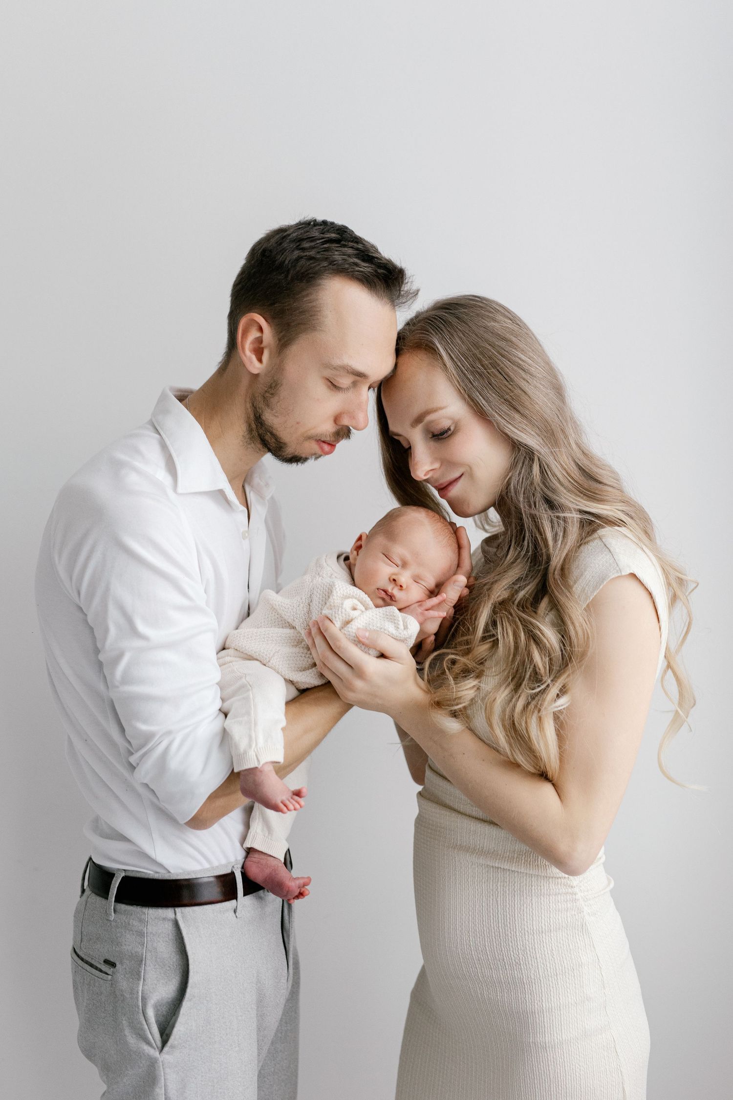 Young parents in white outfits lovingly holding their sleeping newborn baby together.