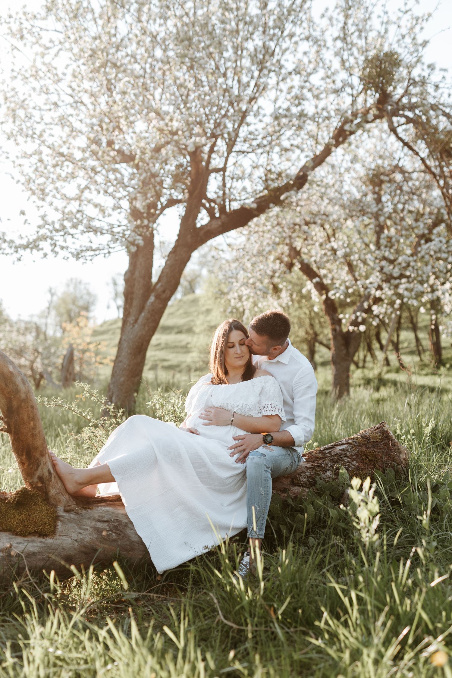 Couple sitting on a fallen log in a blooming spring orchard, man kissing woman in white dress.