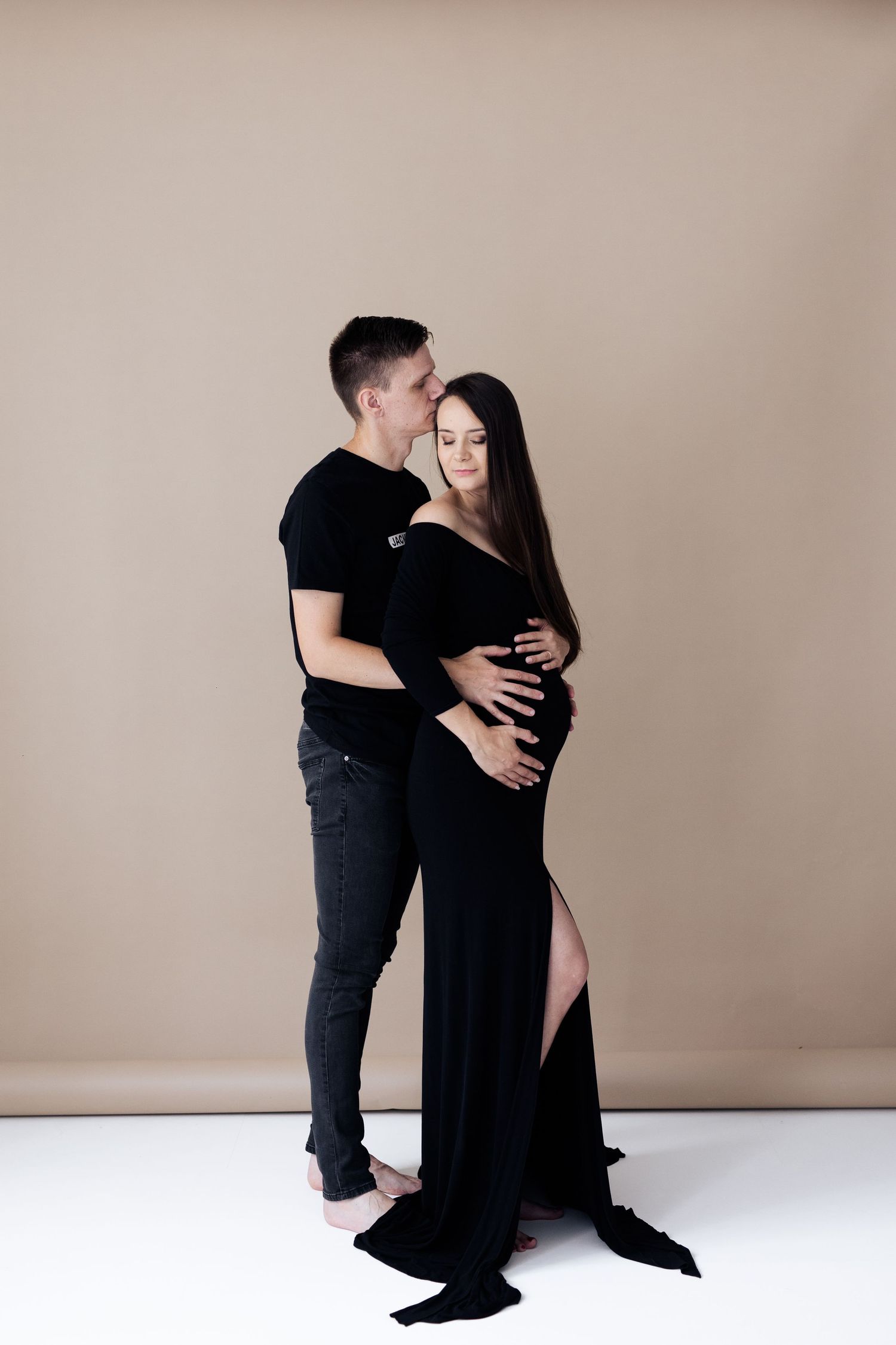 Couple maternity photo with man kissing pregnant woman in black gown in studio.