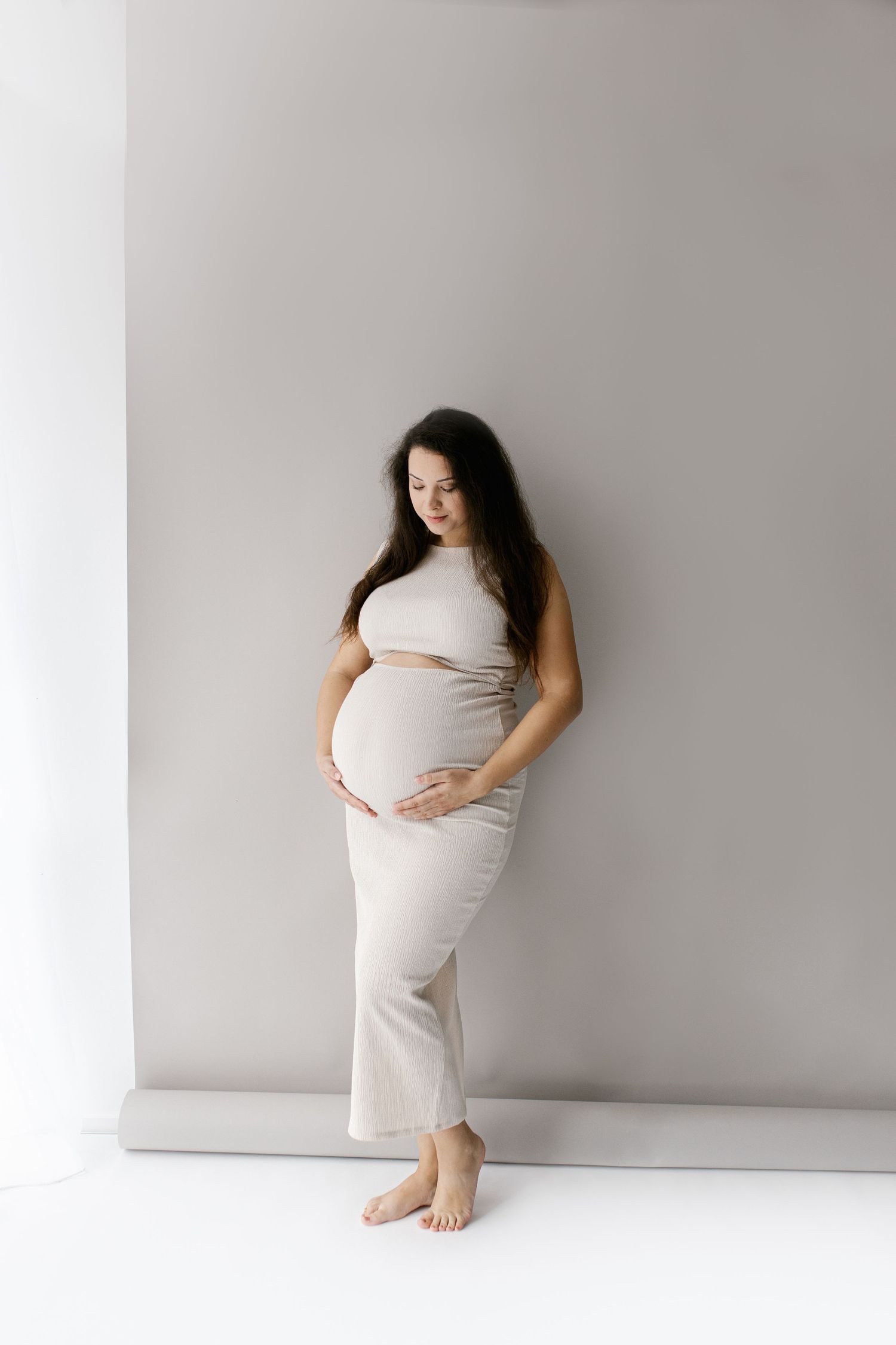Elegant pregnant woman in beige outfit standing against white wall gently holding her baby bump.