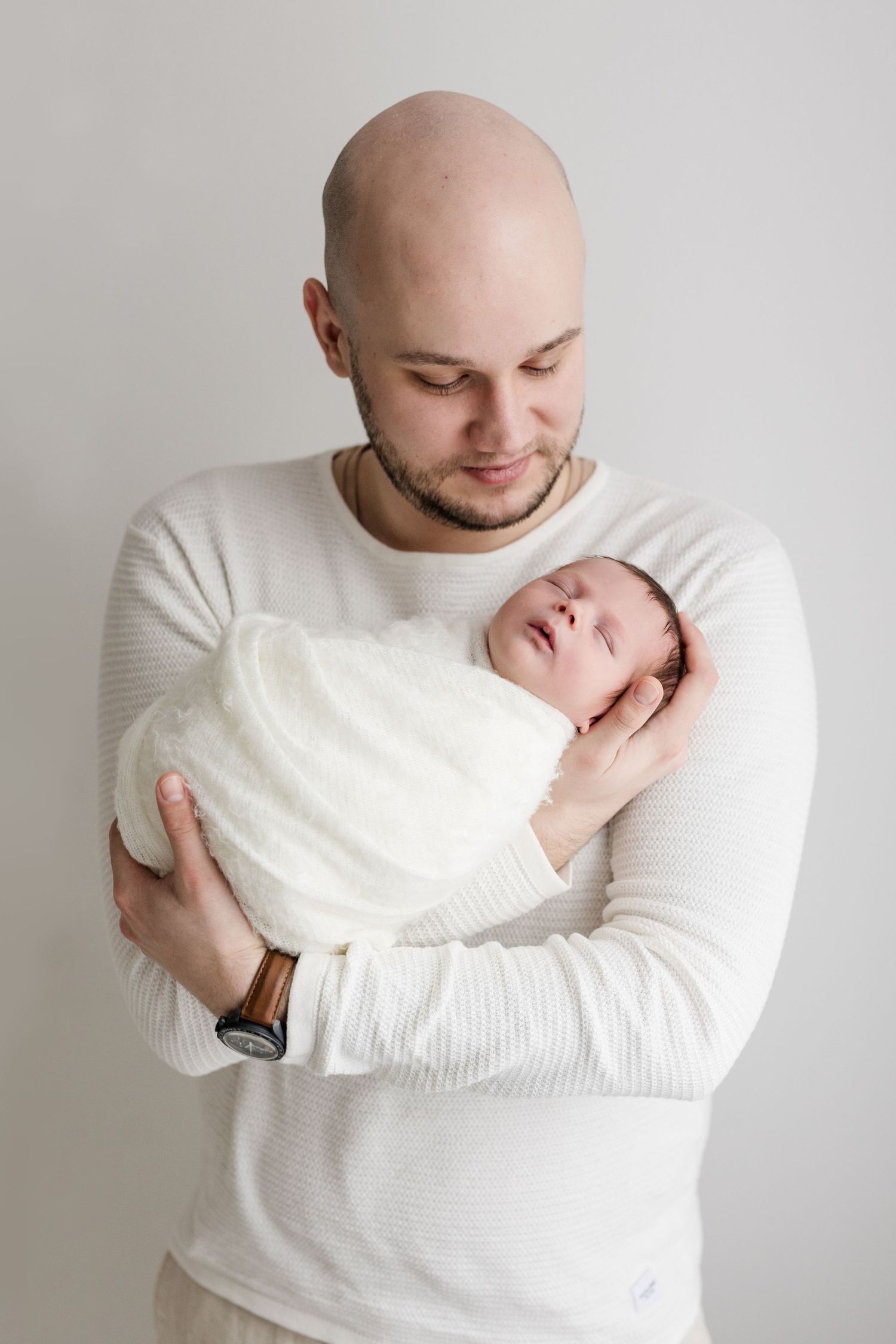 Bald father in white sweater gazes lovingly at his sleeping swaddled newborn baby in studio portrait.