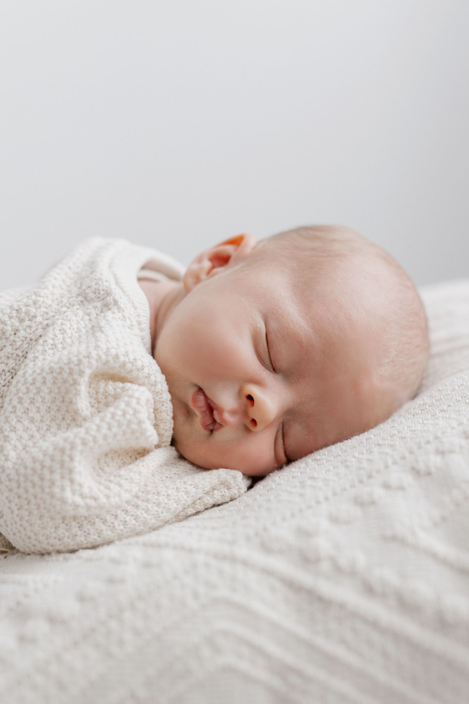 Peaceful newborn baby sleeping wrapped in soft white knit blanket on white surface.