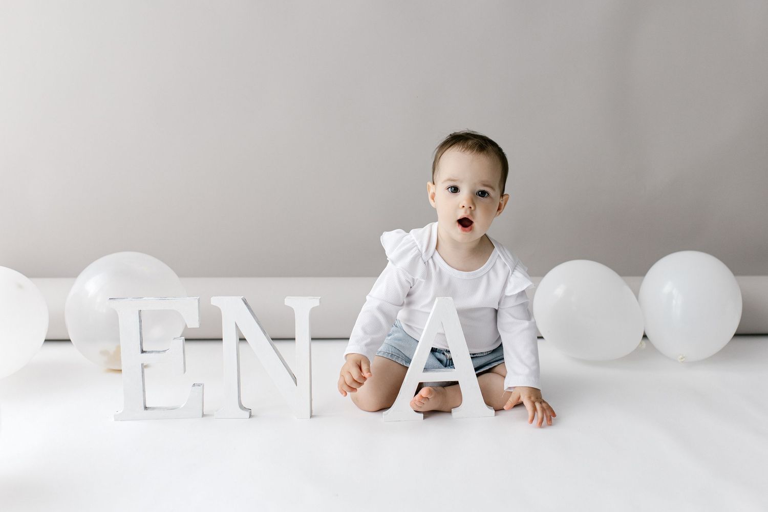 Curious baby boy sitting next to ENA letters with white balloons on white floor.