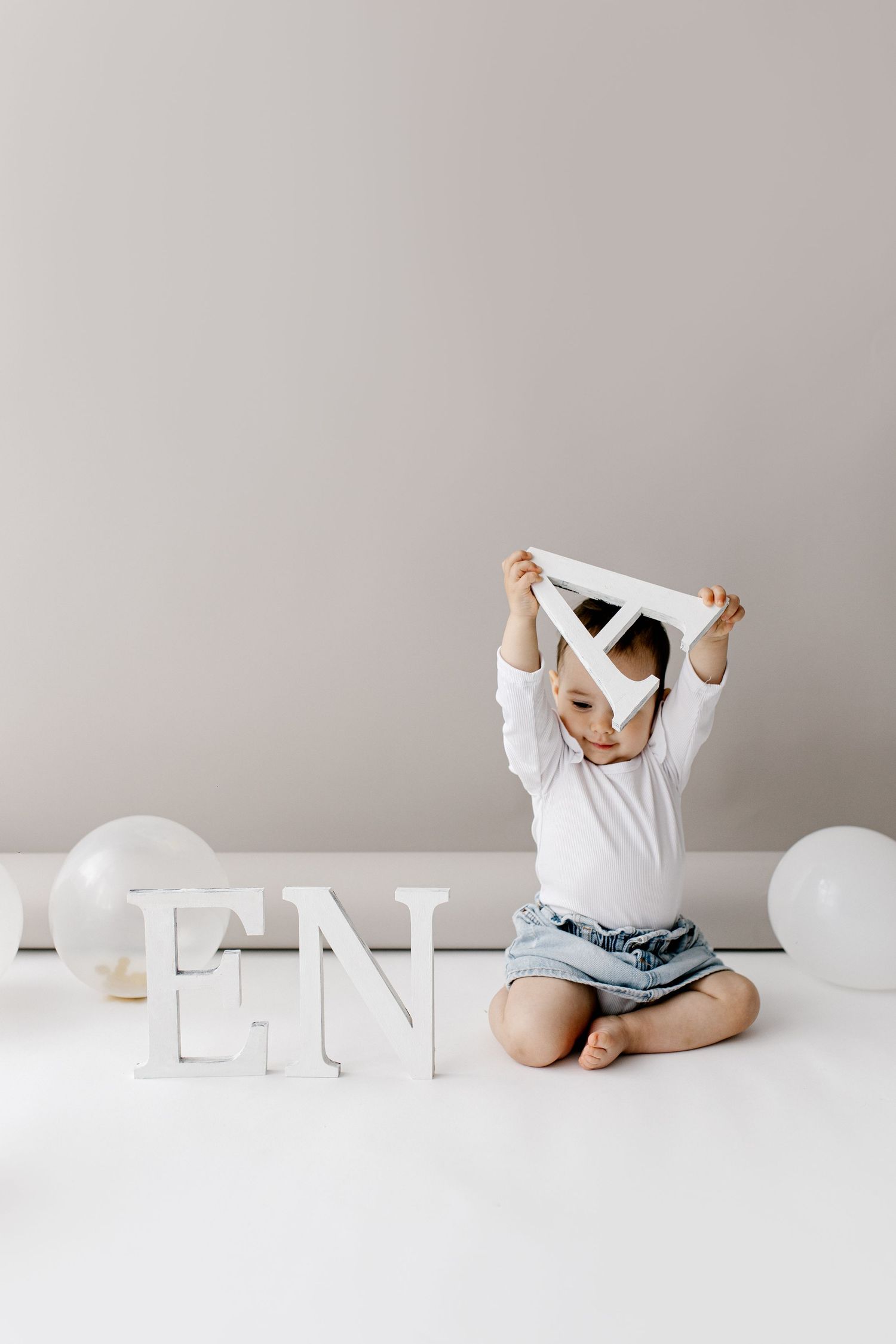 Toddler sitting on floor playing with white letter A next to EN letters and balloons.