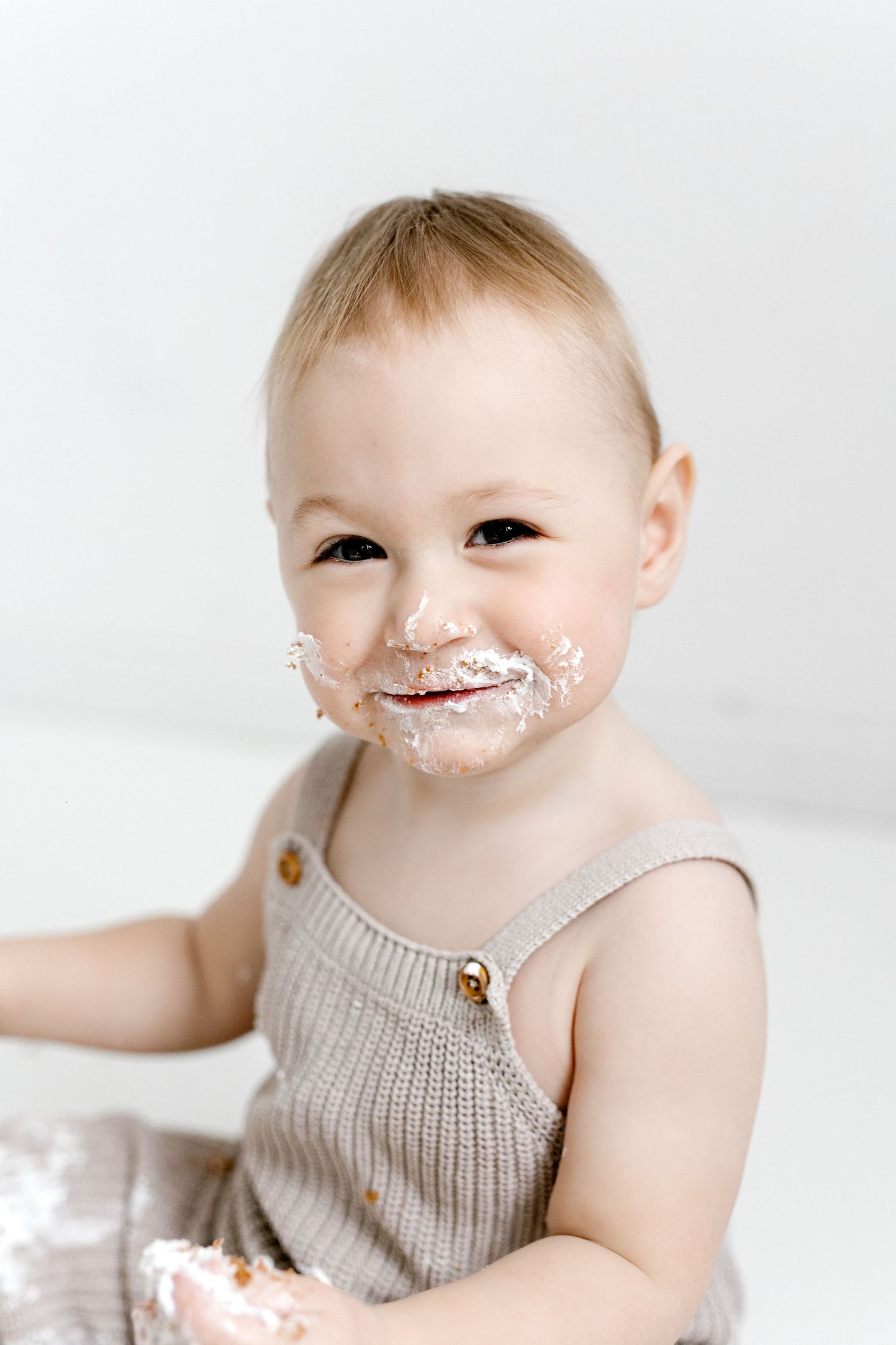 Smiling baby with frosting on face during first birthday cake smash in knit romper.