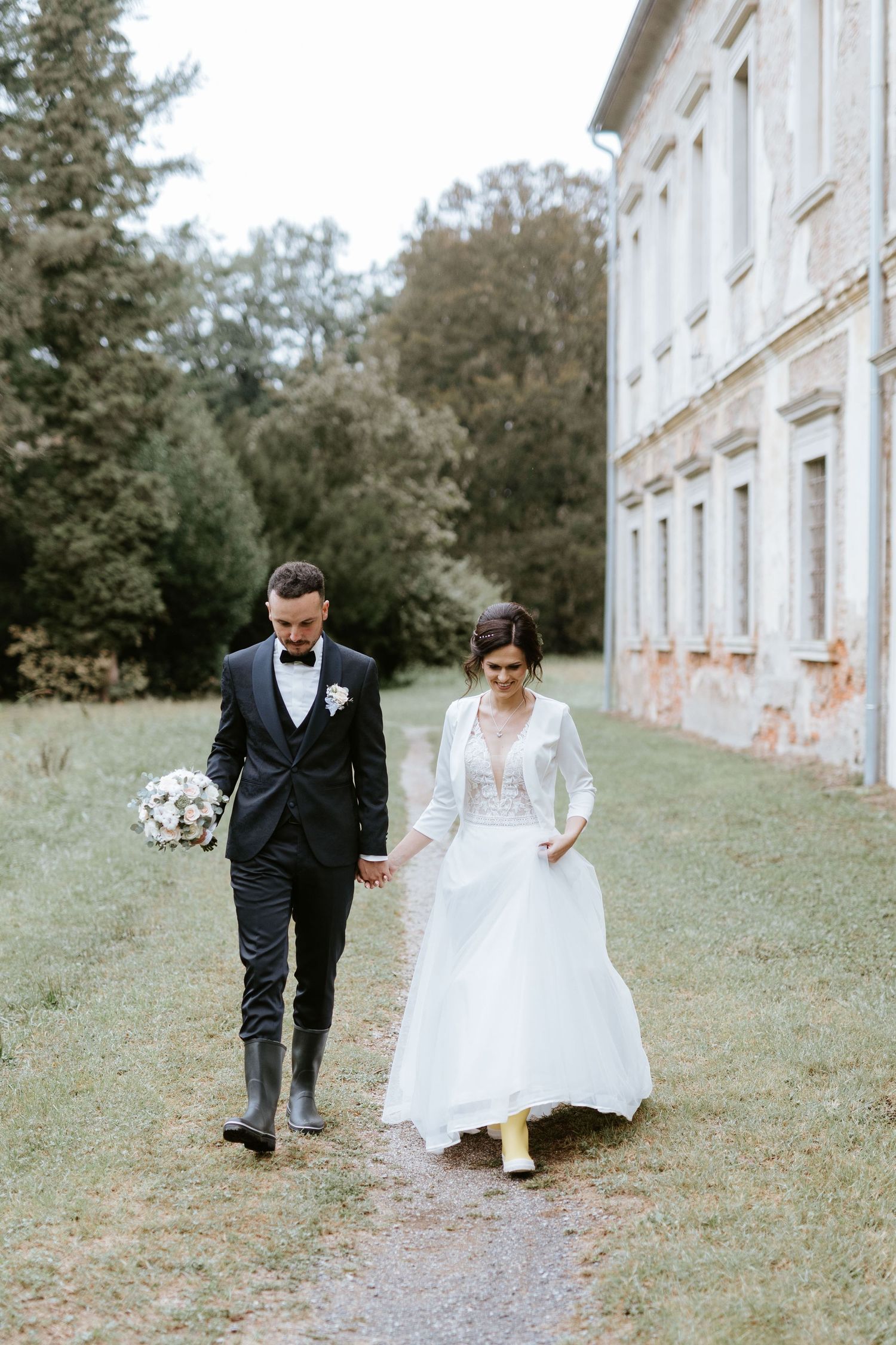 Bride and groom walk hand in hand wearing rain boots past a historic manor house.