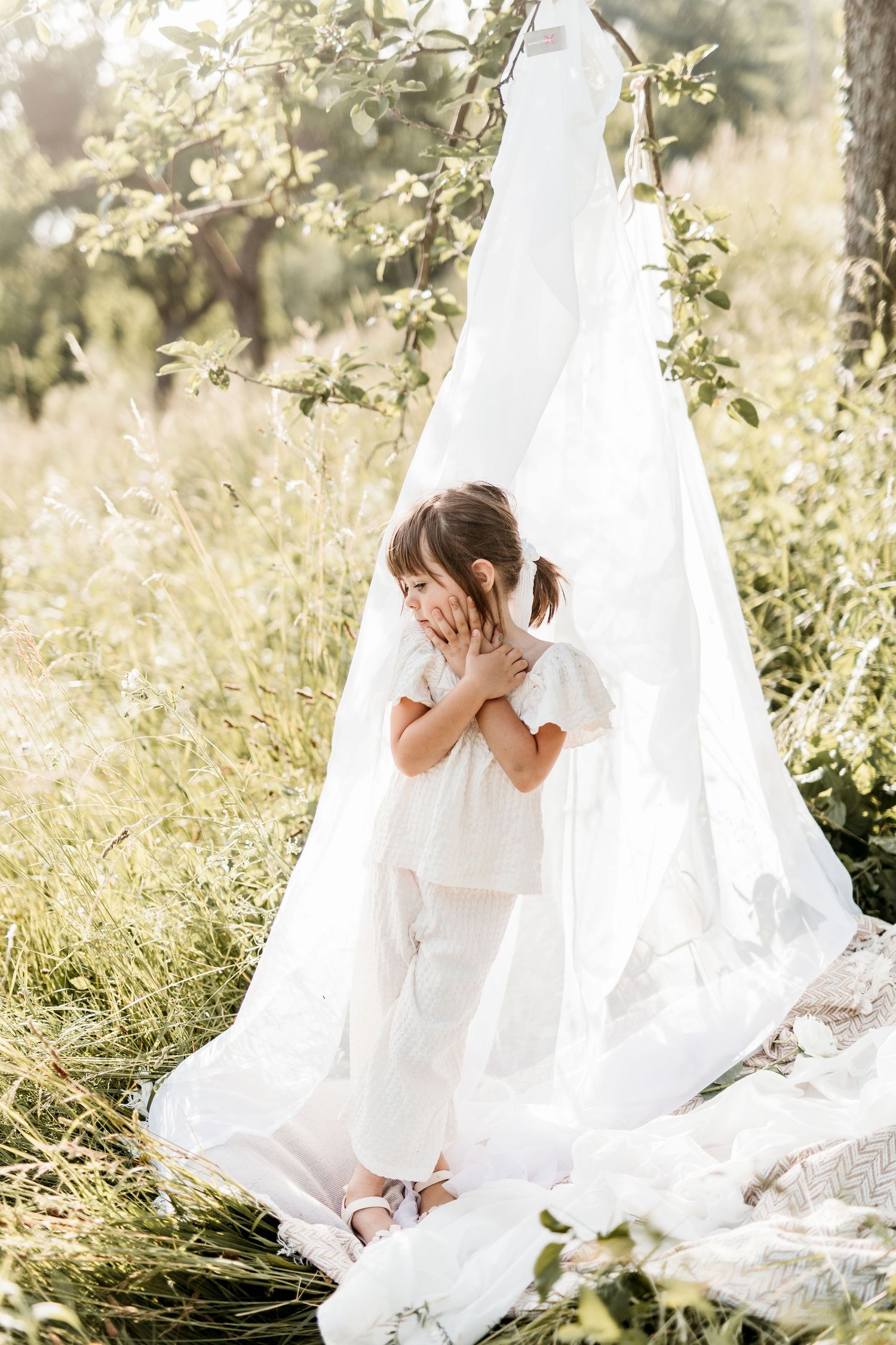 Little girl in white outfit standing under white fabric canopy in nature.