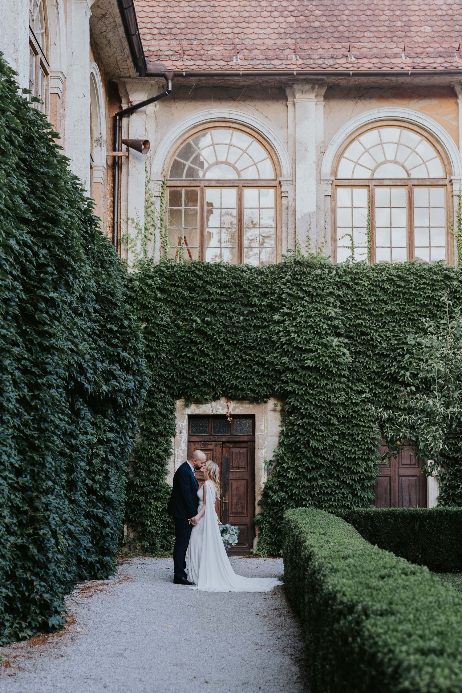Bride and groom share a romantic kiss in a lush ivy-covered European estate garden with arched windows.