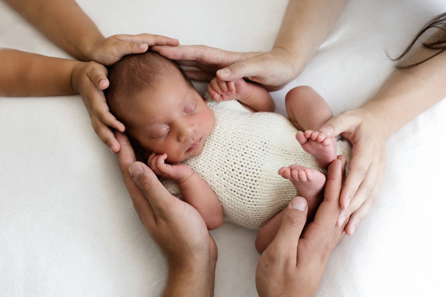 Sleeping newborn baby in knit wrap cradled by four loving parents hands.