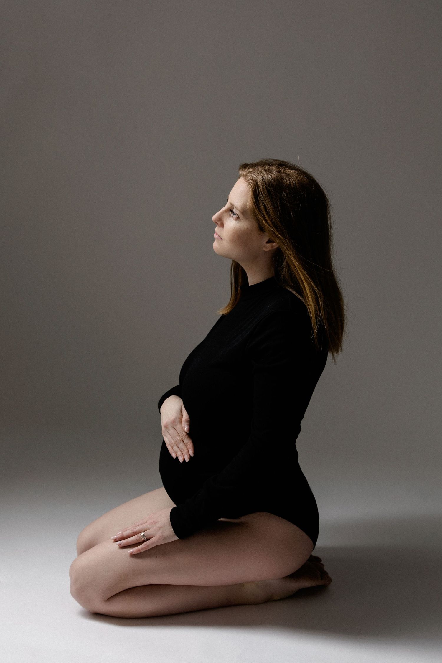 Pregnant woman in black bodysuit sitting on floor in profile view on gray background.
