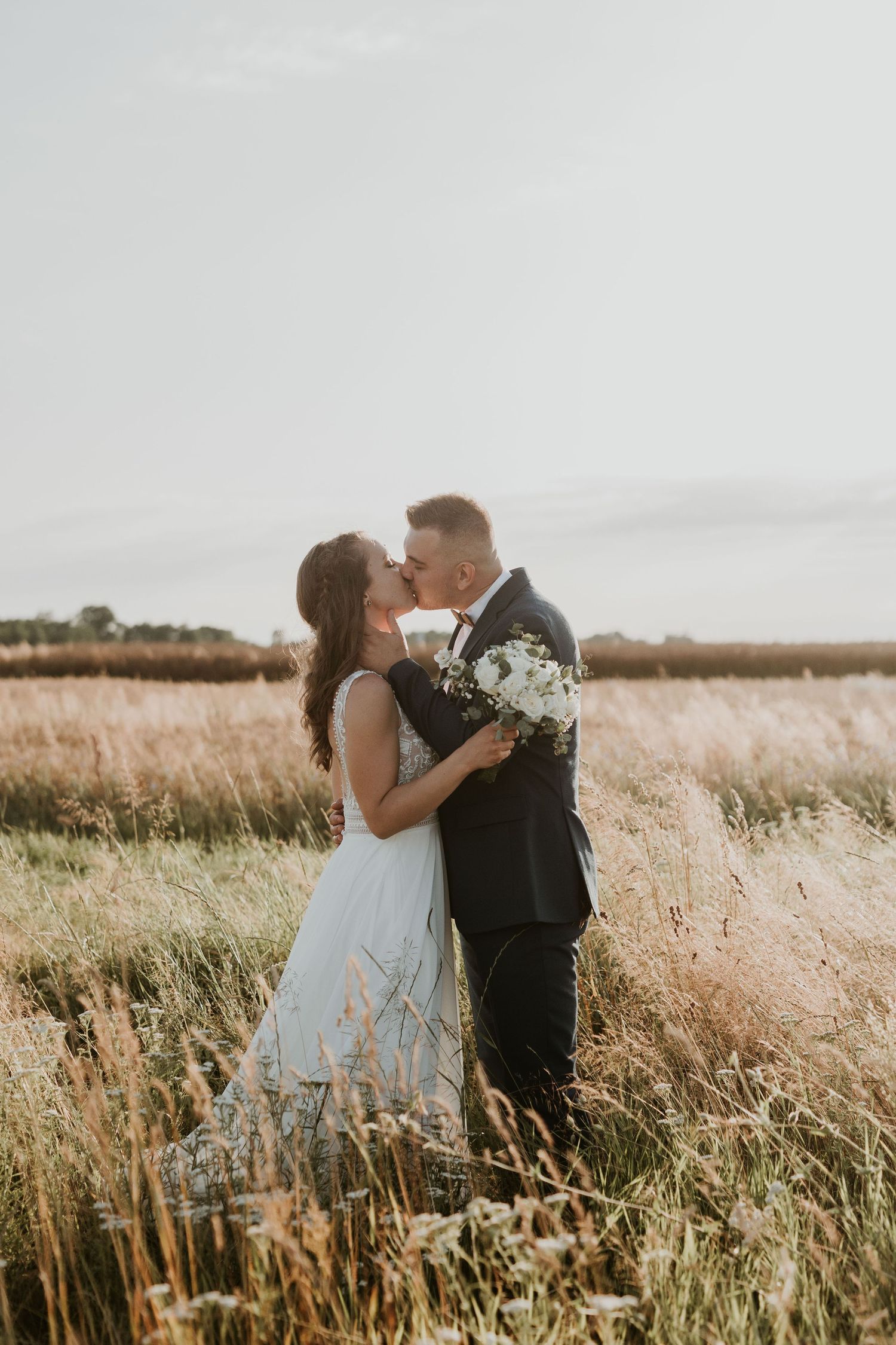Bride and groom share a romantic kiss in a golden wheat field under an open sky.