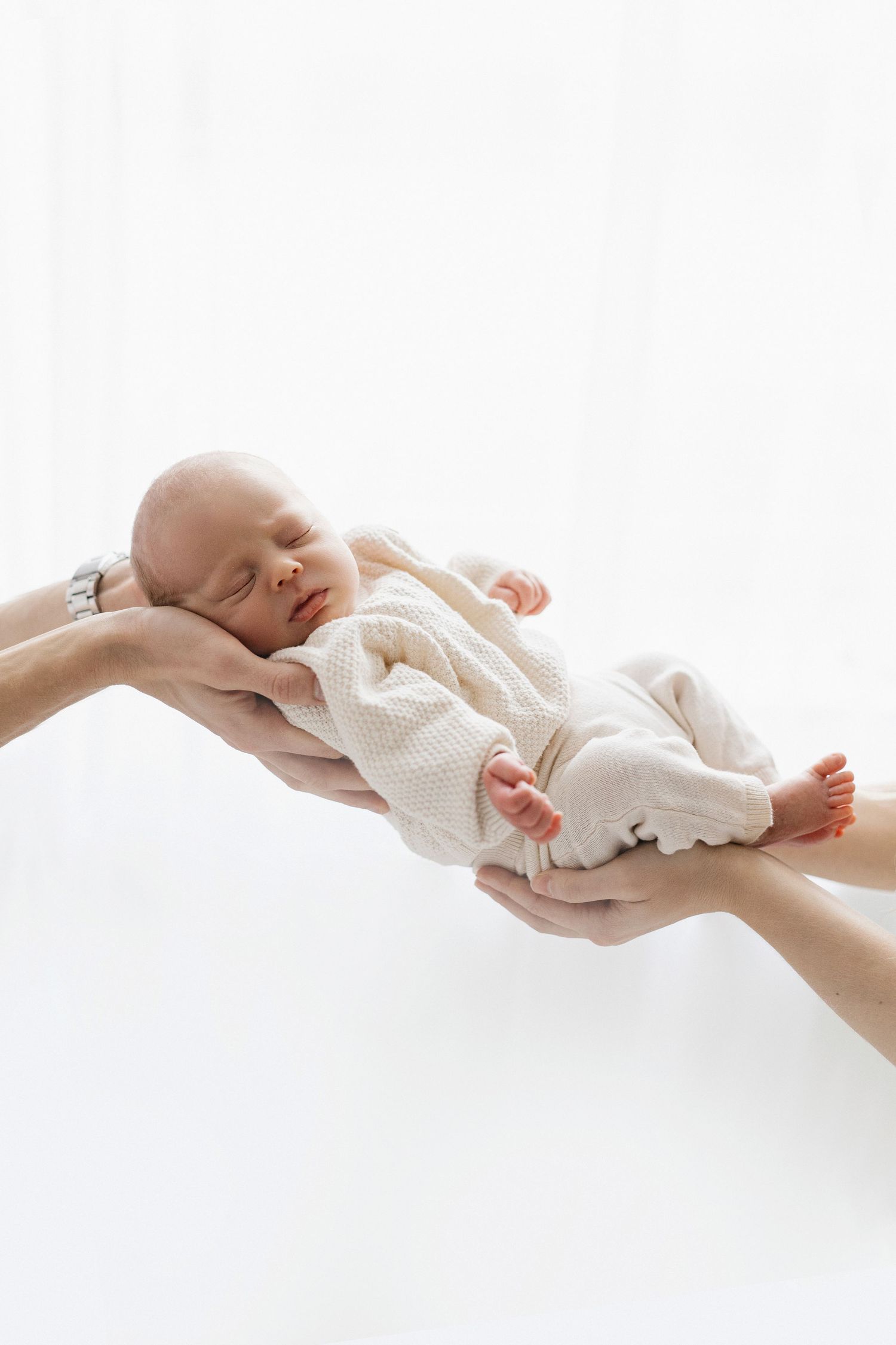 Parents gently holding sleeping newborn baby in white onesie against bright white background.