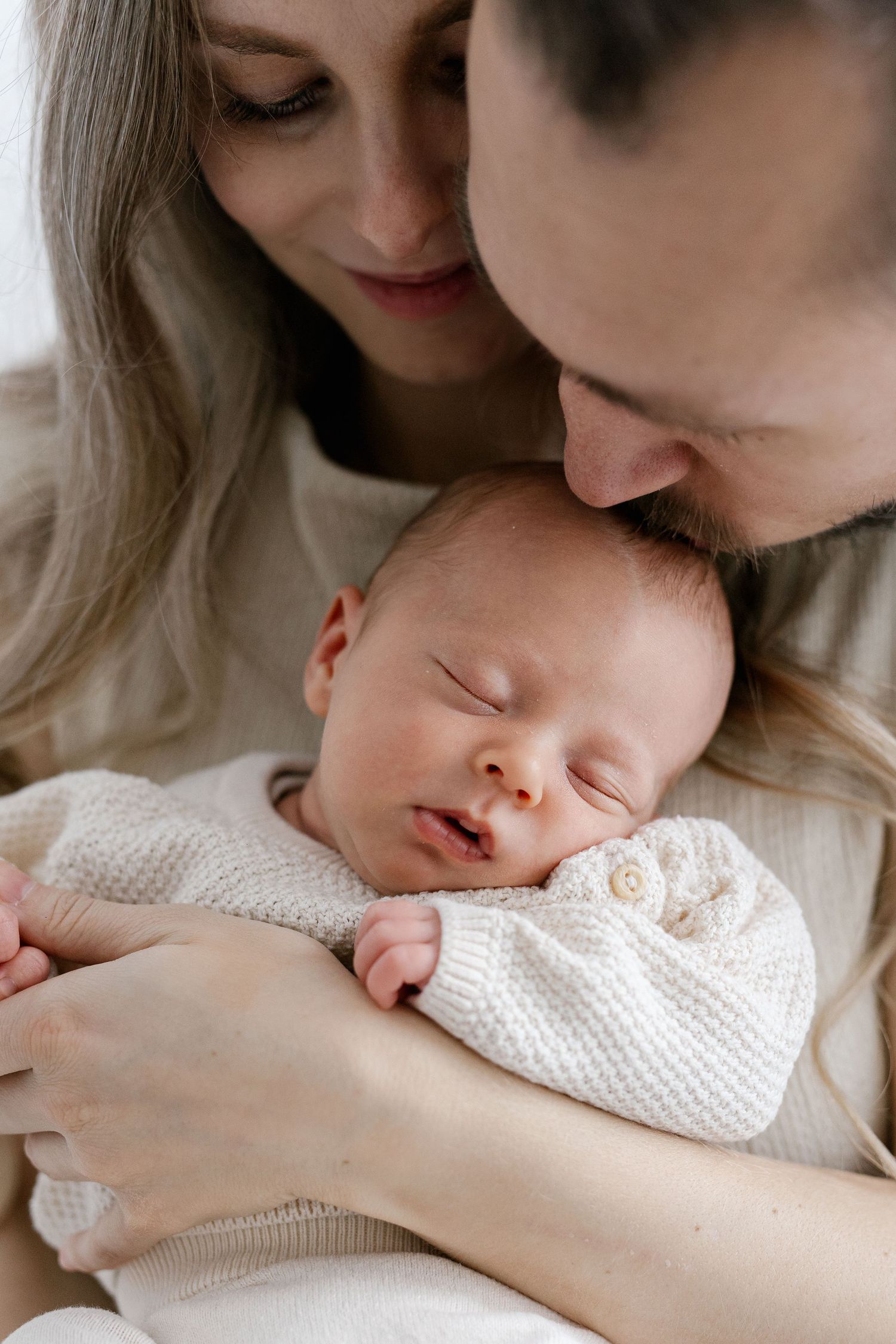 Parents tenderly kissing their sleeping newborn baby wrapped in a white knit outfit.