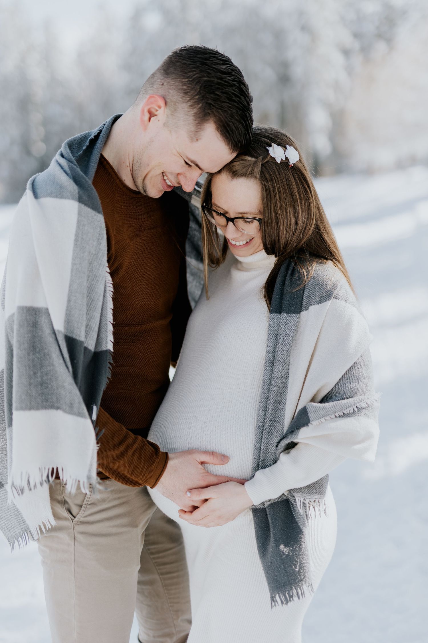 Smiling couple sharing tender moment, hands on pregnant belly in snowy winter landscape.