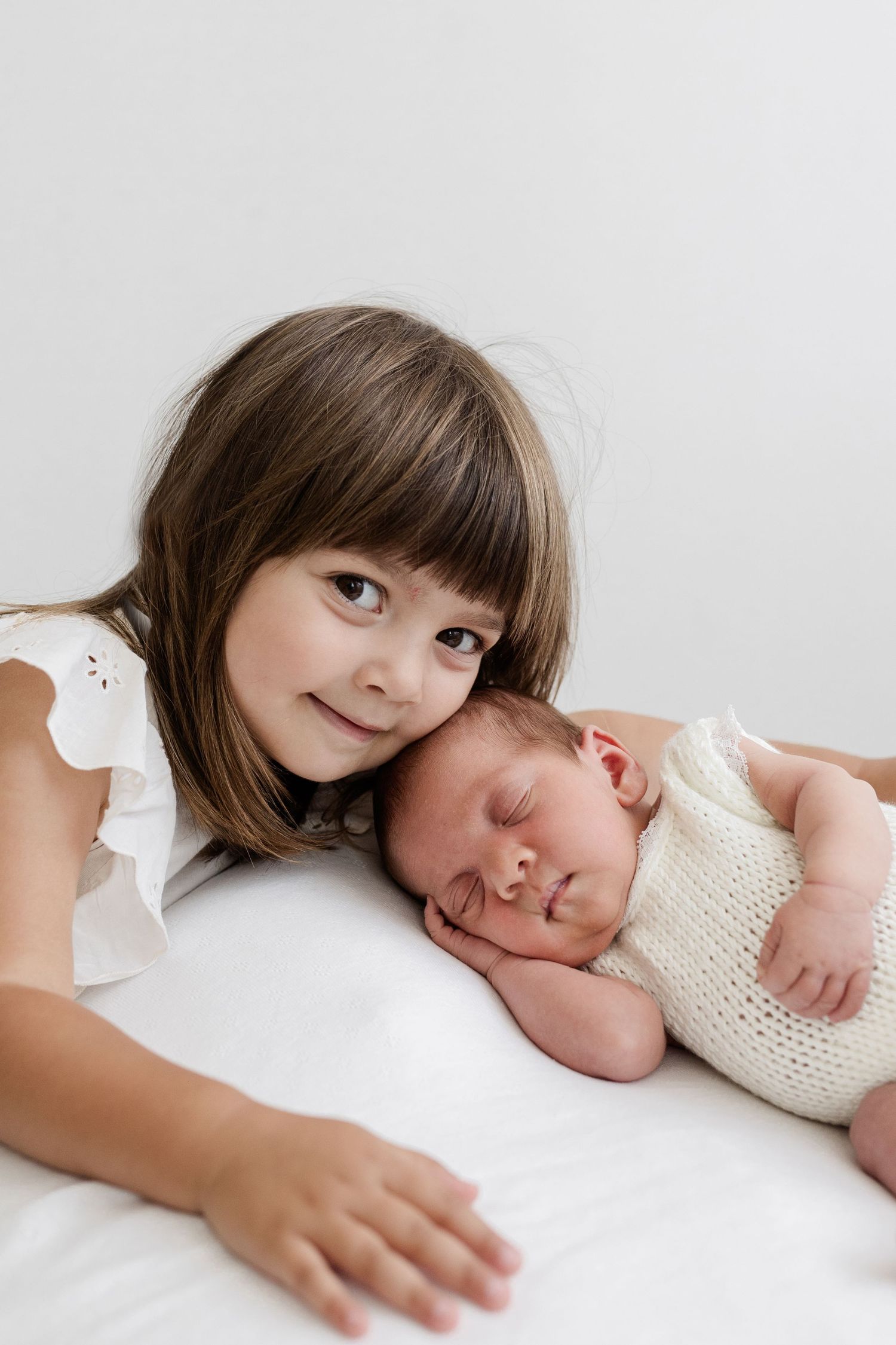 A smiling young girl with brown hair poses cheek-to-cheek next to her sleeping newborn sibling.