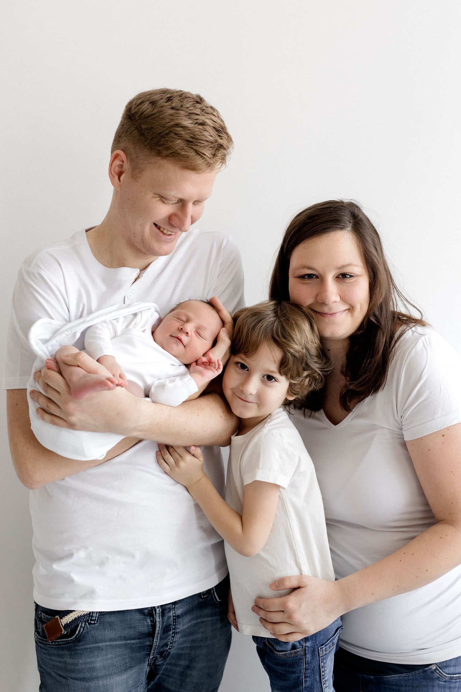 Smiling family of four in white tops and jeans pose together with their newborn baby in a studio.