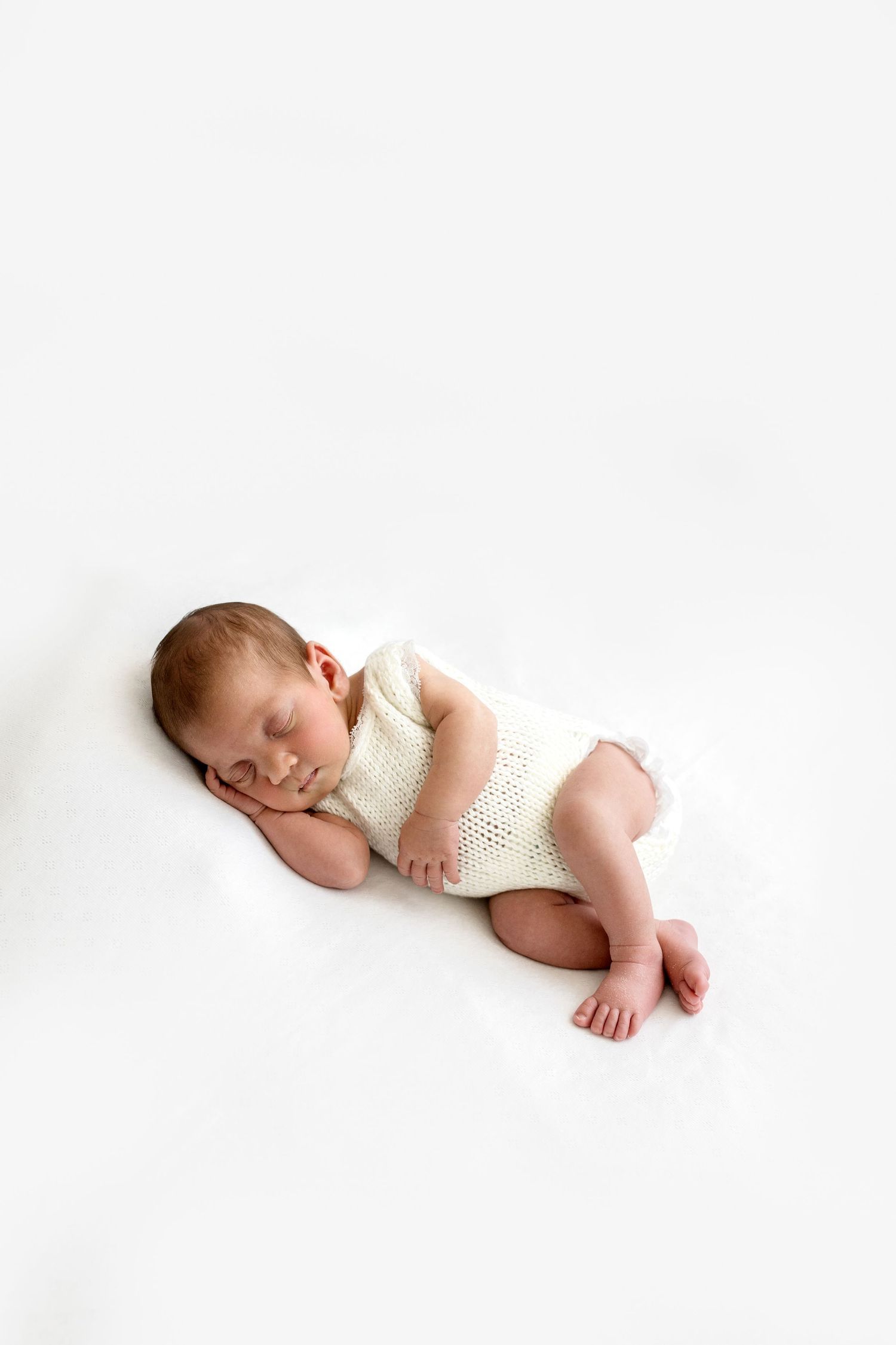 Peaceful newborn baby sleeping curled up in cream knit romper on white background.