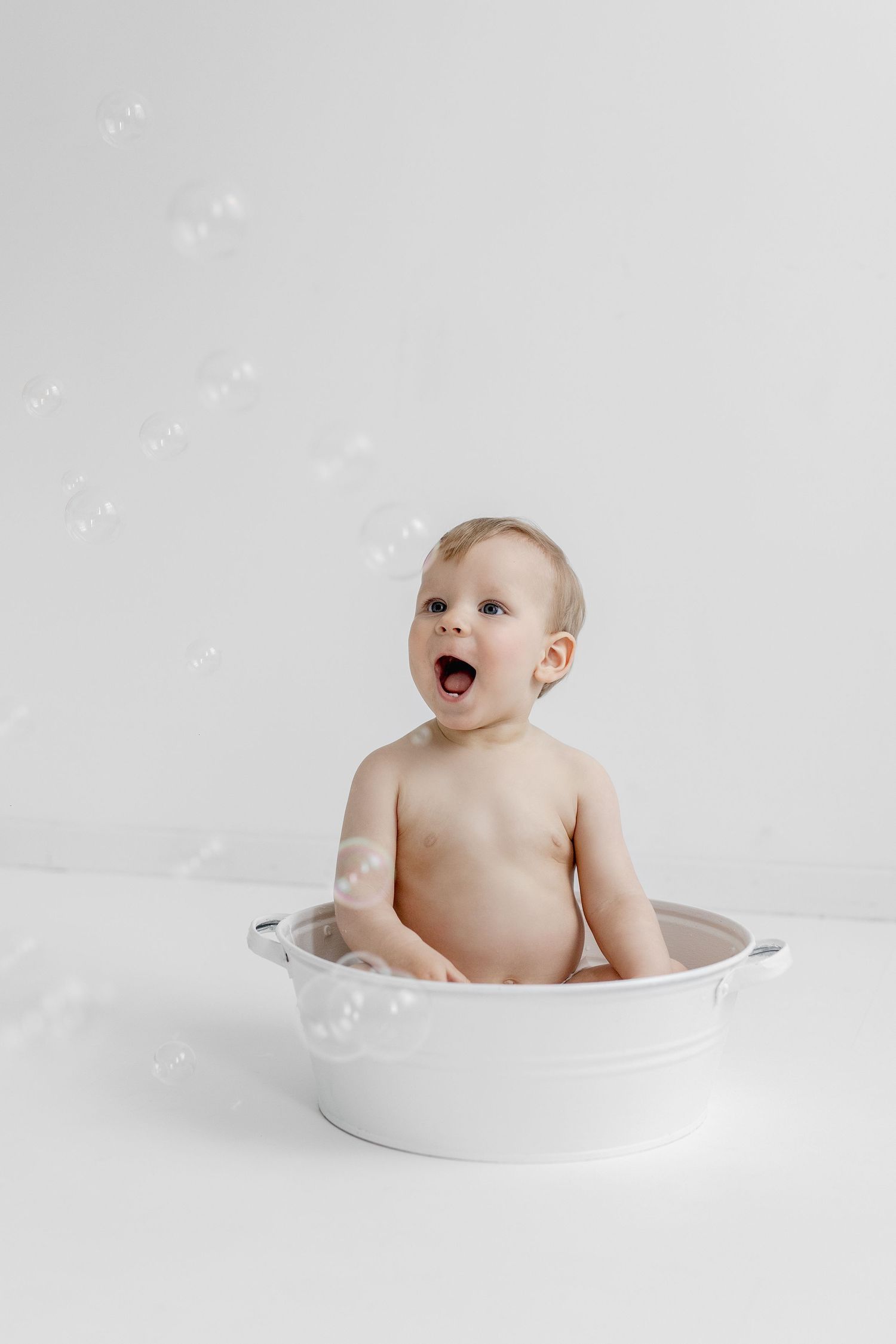 A delighted baby sits in a white basin tub surrounded by floating soap bubbles against a white background.