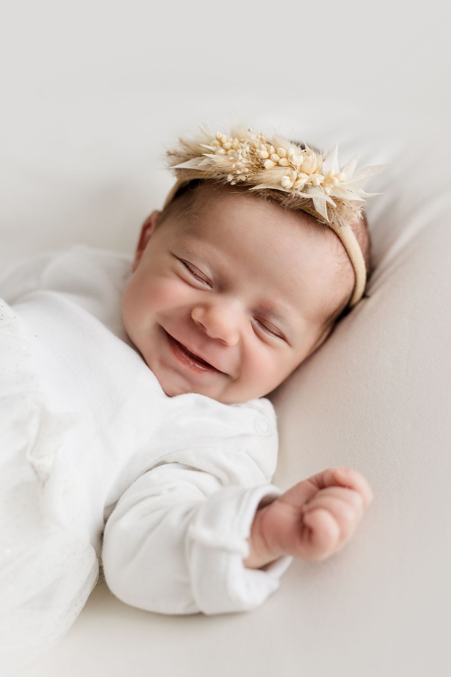 Smiling newborn baby girl wearing a delicate dried flower headband, dressed in white, lying on a soft surface.