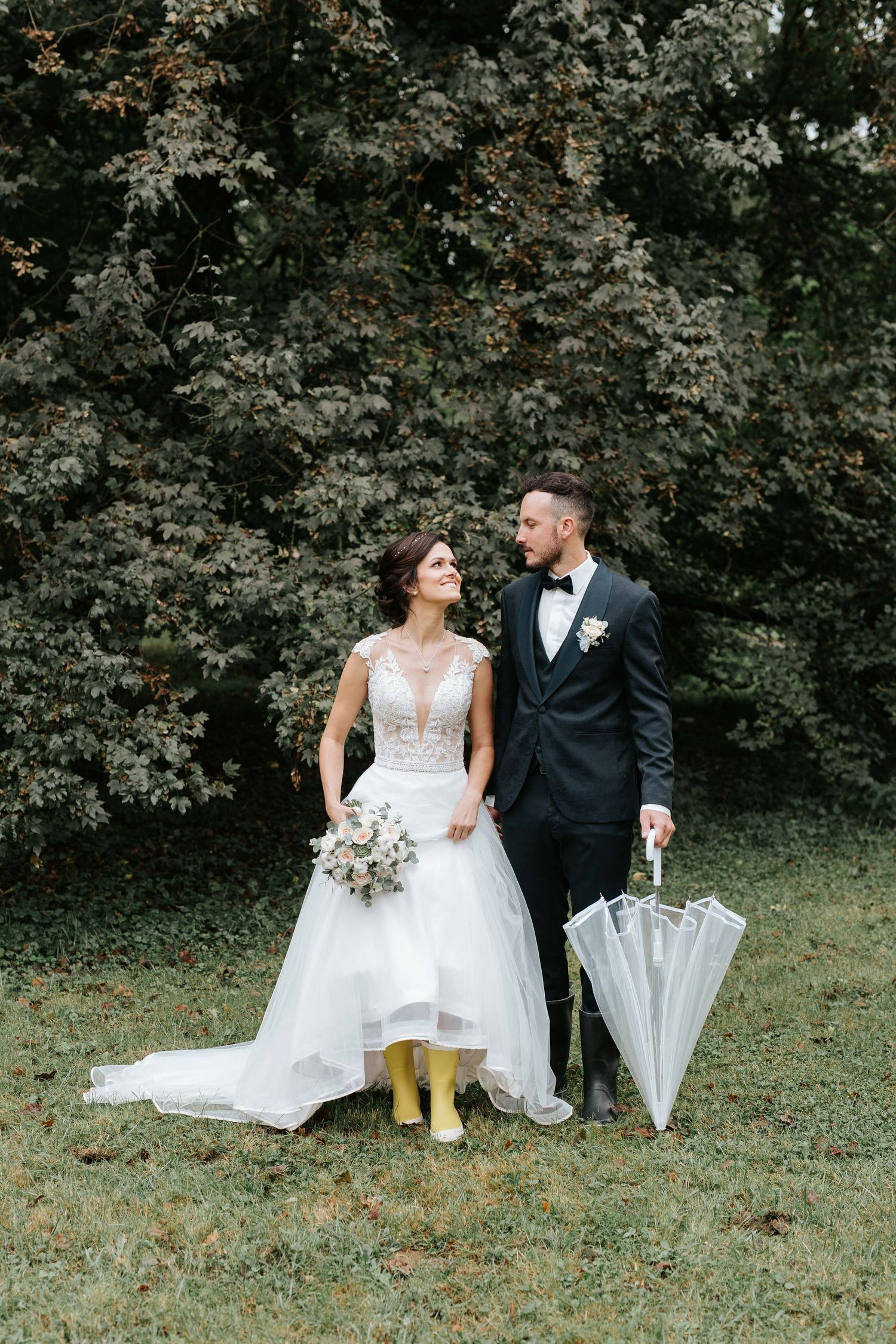 Bride in white dress with yellow rain boots and groom holding clear umbrella standing before lush green trees.