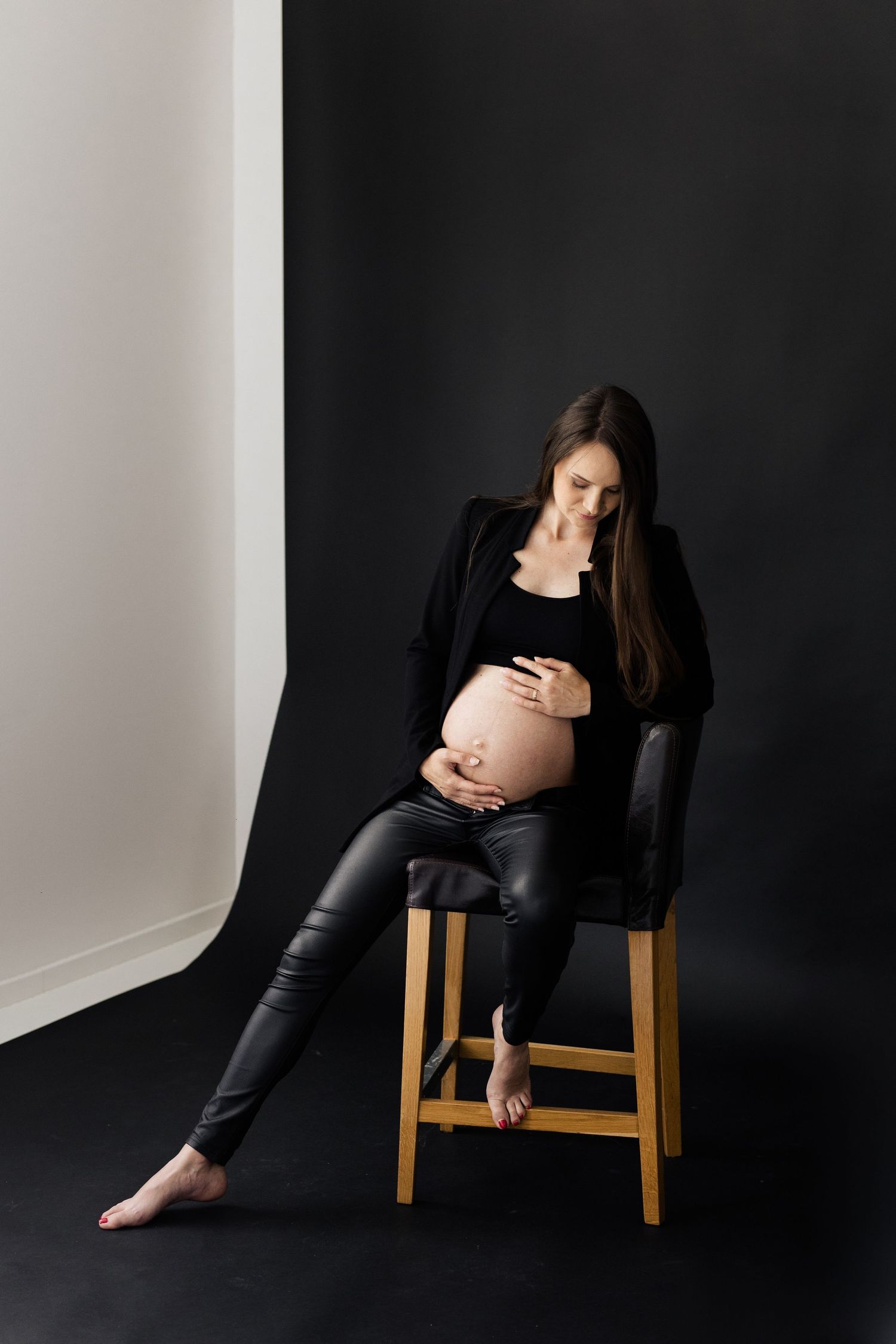 Pregnant woman in black outfit seated on stool holding baby bump in dark studio.