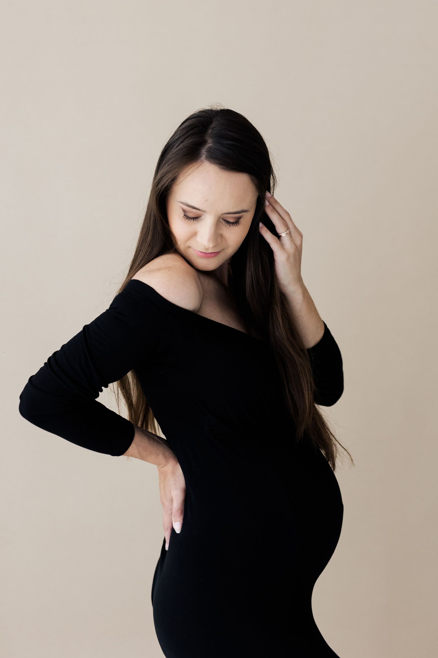 Pregnant woman in black off-shoulder dress cradling her baby bump in a studio.