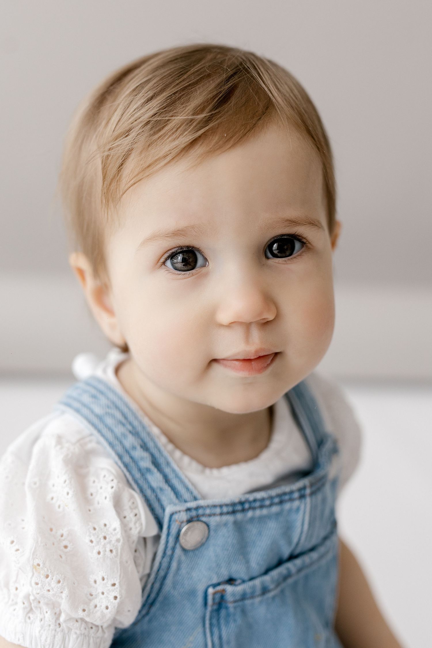 Adorable blue-eyed baby girl in denim overalls and white top smiling at camera.