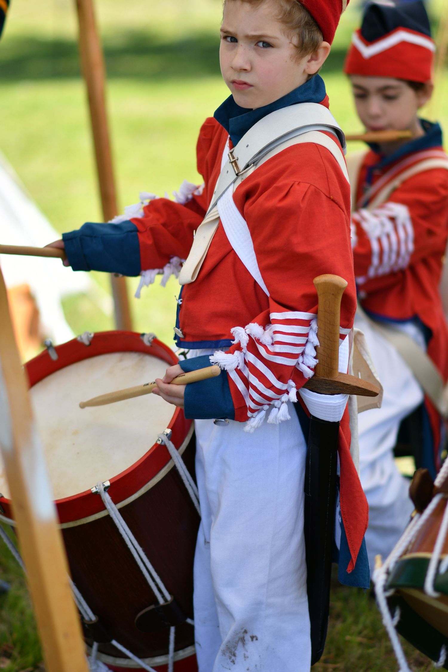 A young drummer in a red military uniform participates in a historical reenactment.