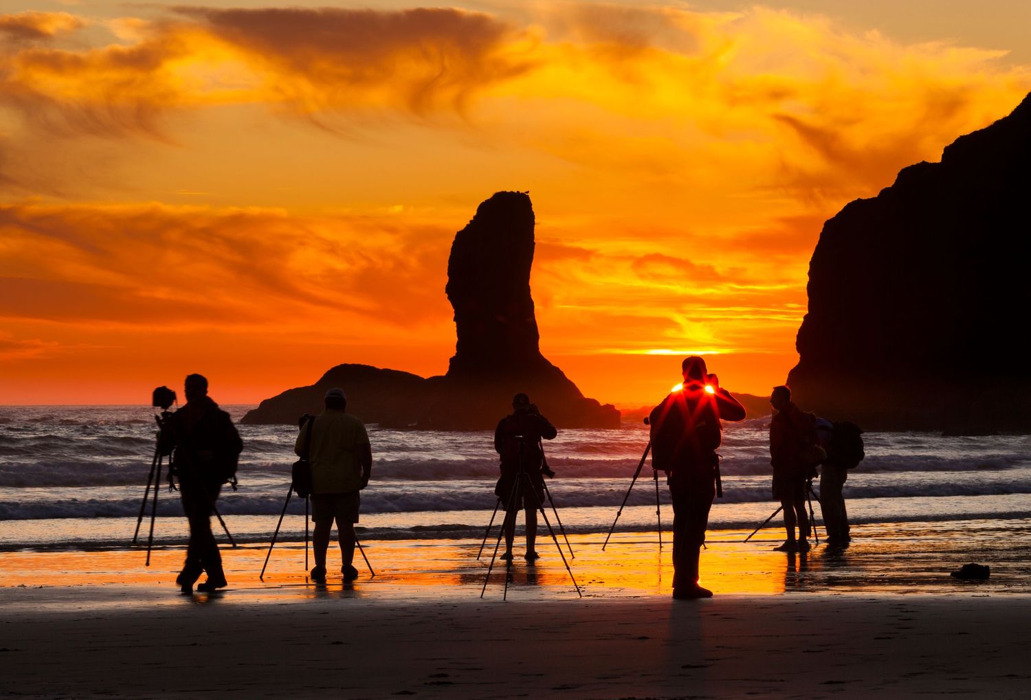Silhouetted photographers with cameras on tripods capture a dramatic orange sunset at a beach with rock formations.