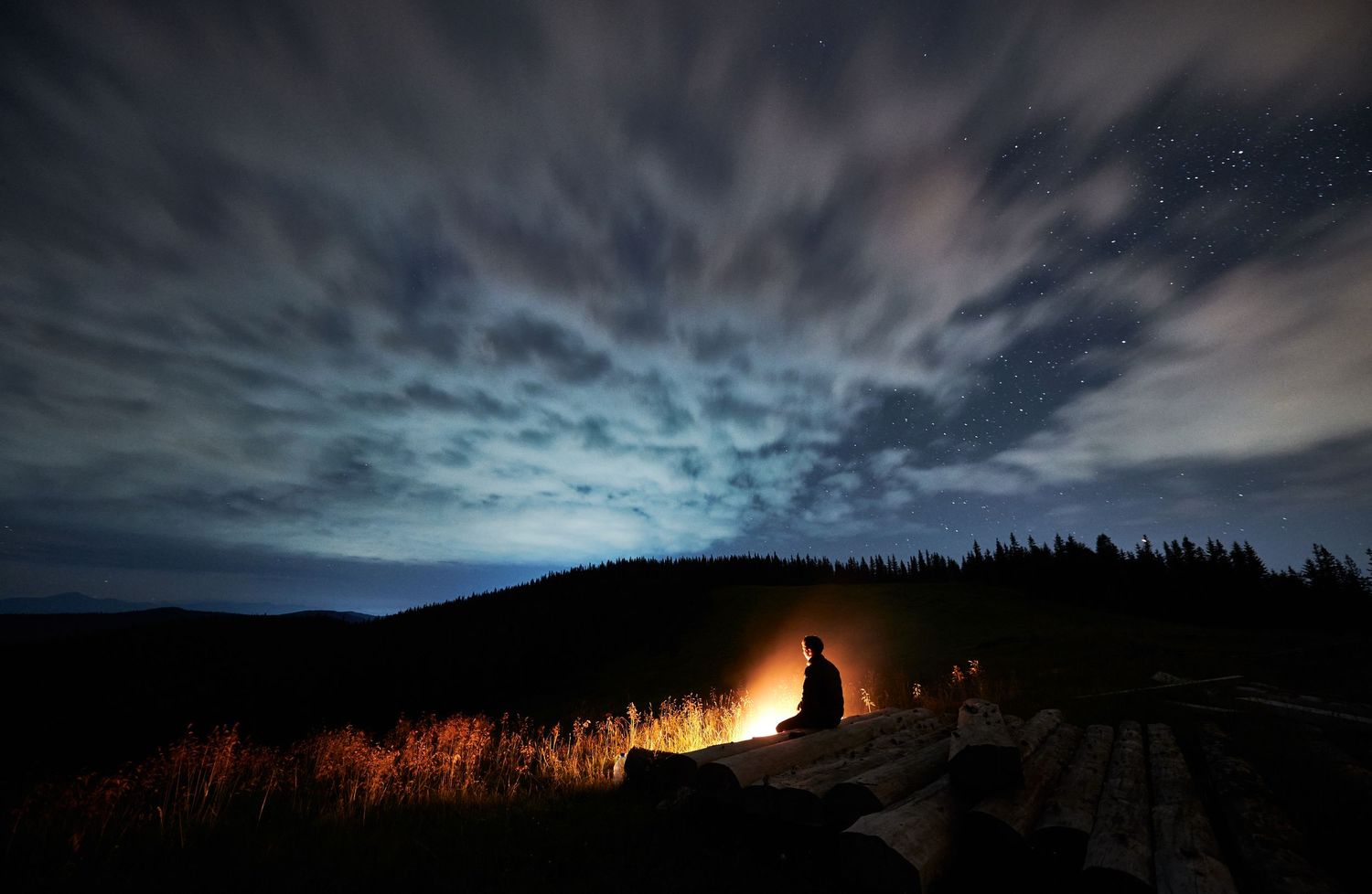Long exposure night photography captures a bright campfire against silhouetted trees and a dramatic cloudy sky.
