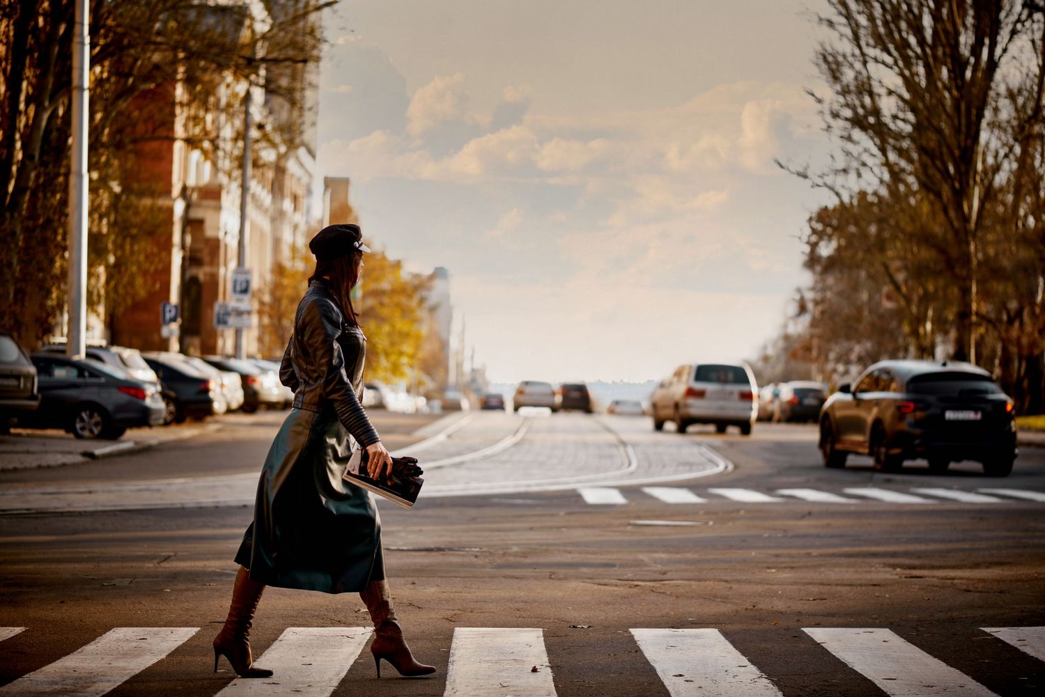 A person in a long dress crosses an urban street at sunset while carrying a bag.