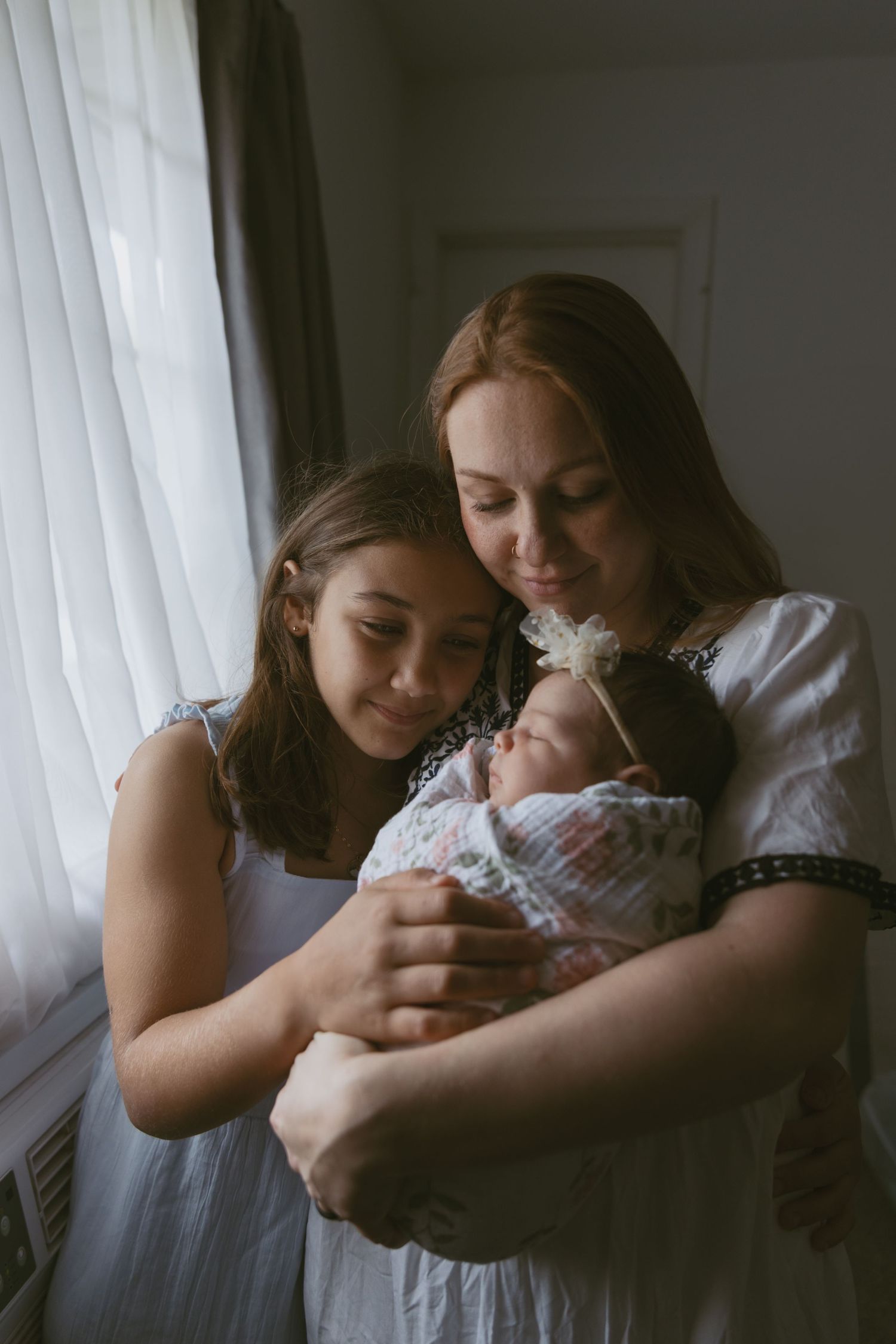 A mom and daughter holding newborn baby next to the window during an in-home photoshoot in Sussex County New Jersey