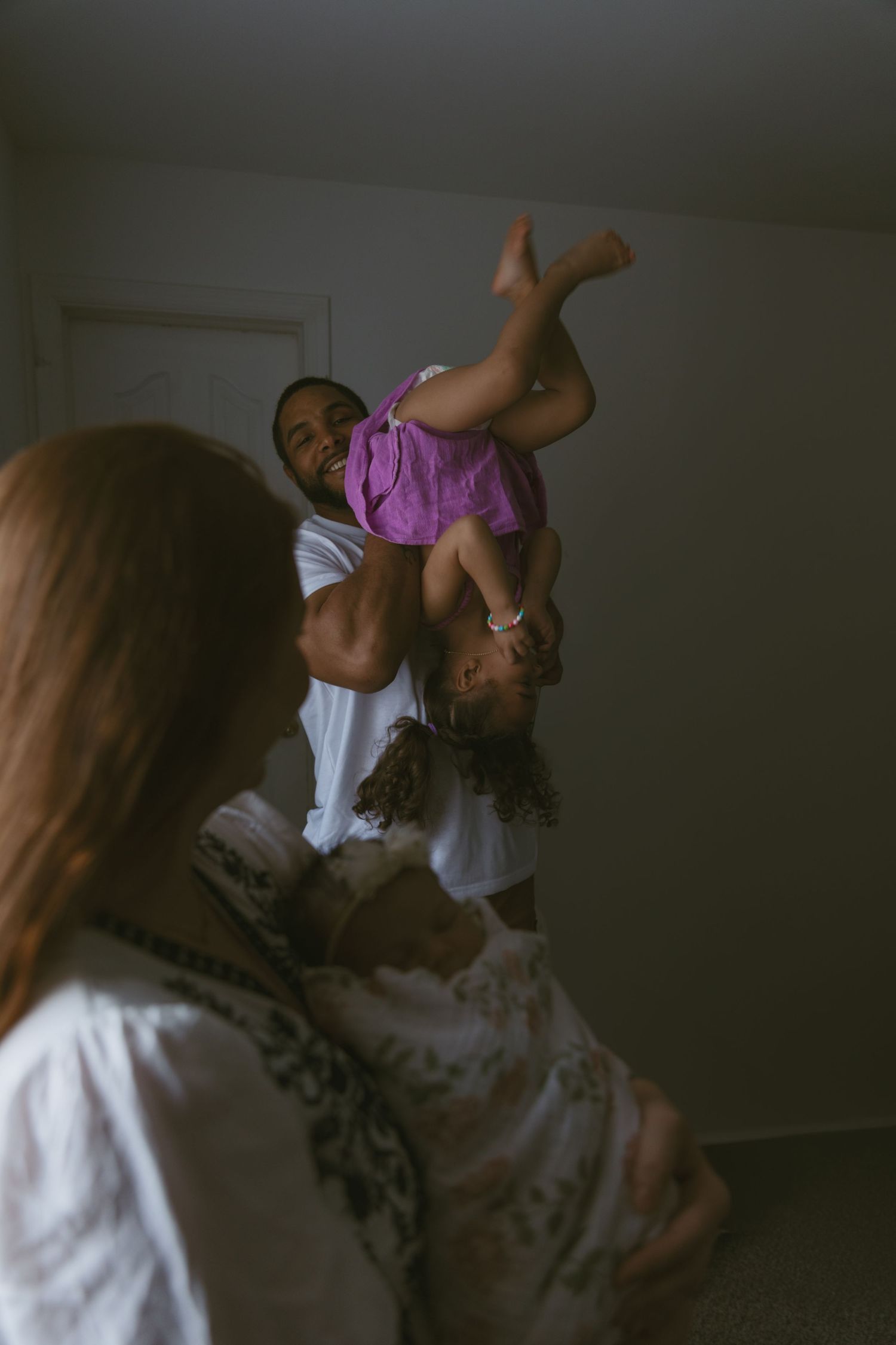 A candid moment of dad playing with toddler, as mom watches while she cradles her newborn baby.  An in-home photoshoot in Sussex New Jersey