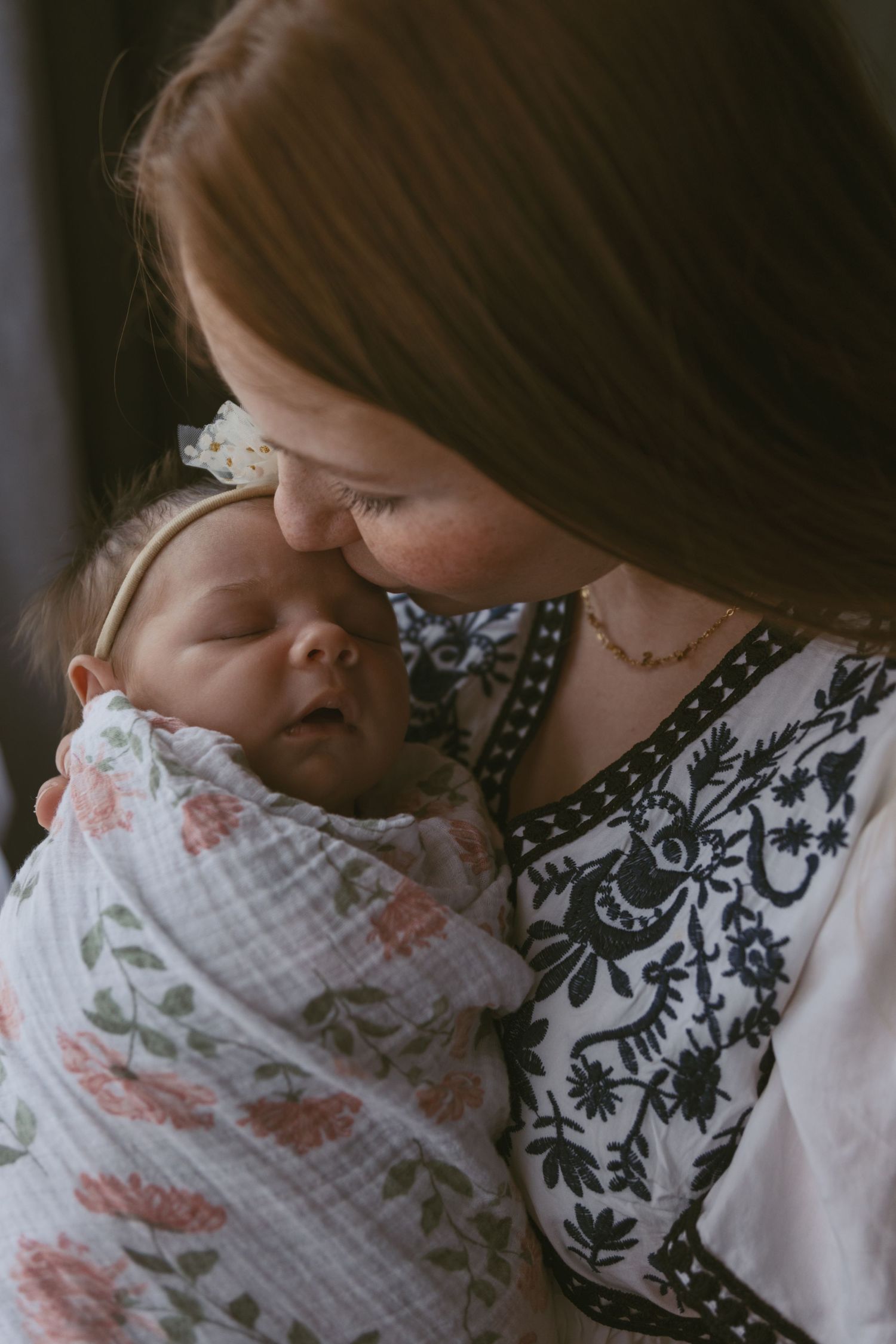 Mother kissing her newborn baby next to the window during an in-home photoshoot in Sussex, New Jersey.