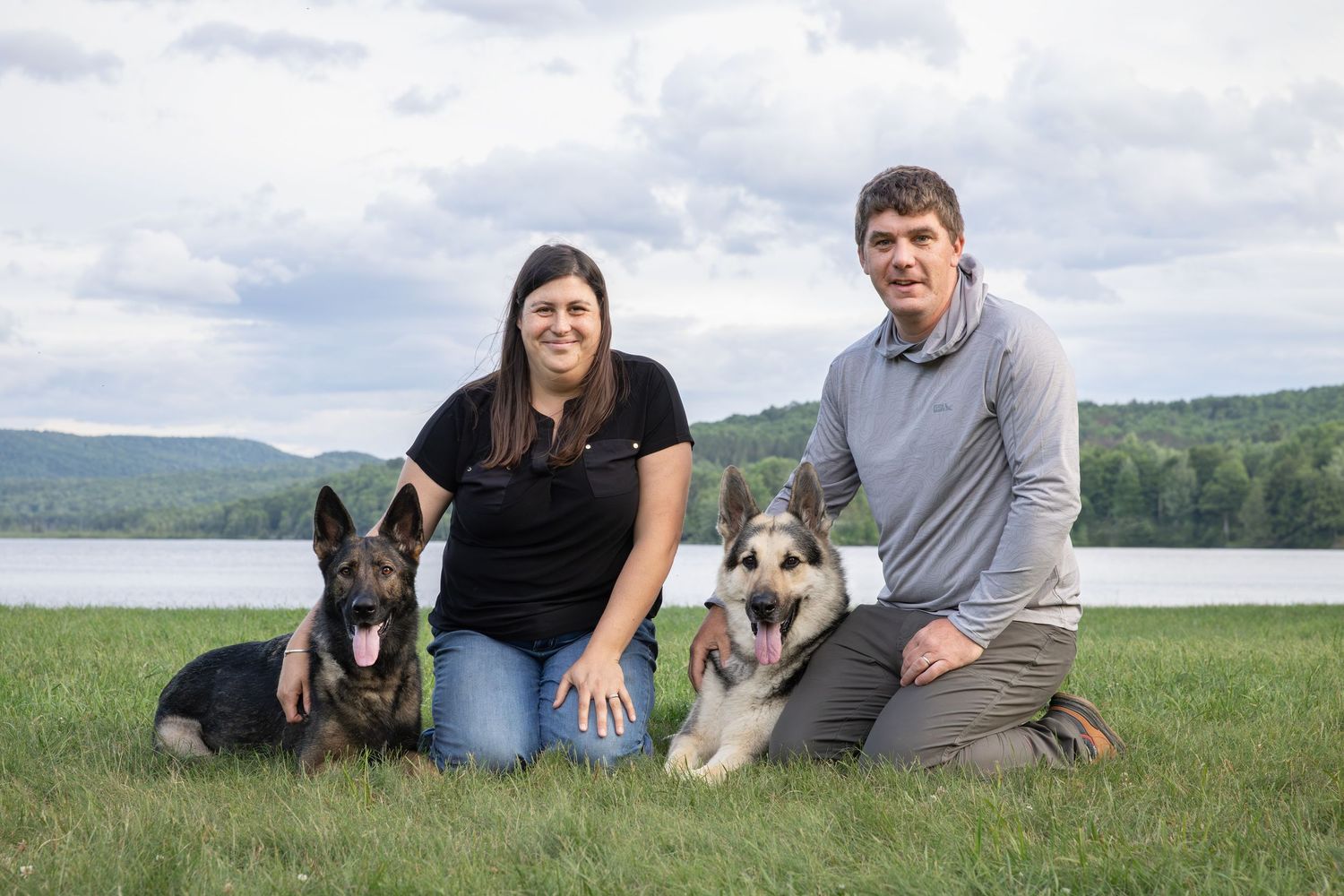 Couple posing outdoors with two German Shepherd dogs on green grass near a lake.