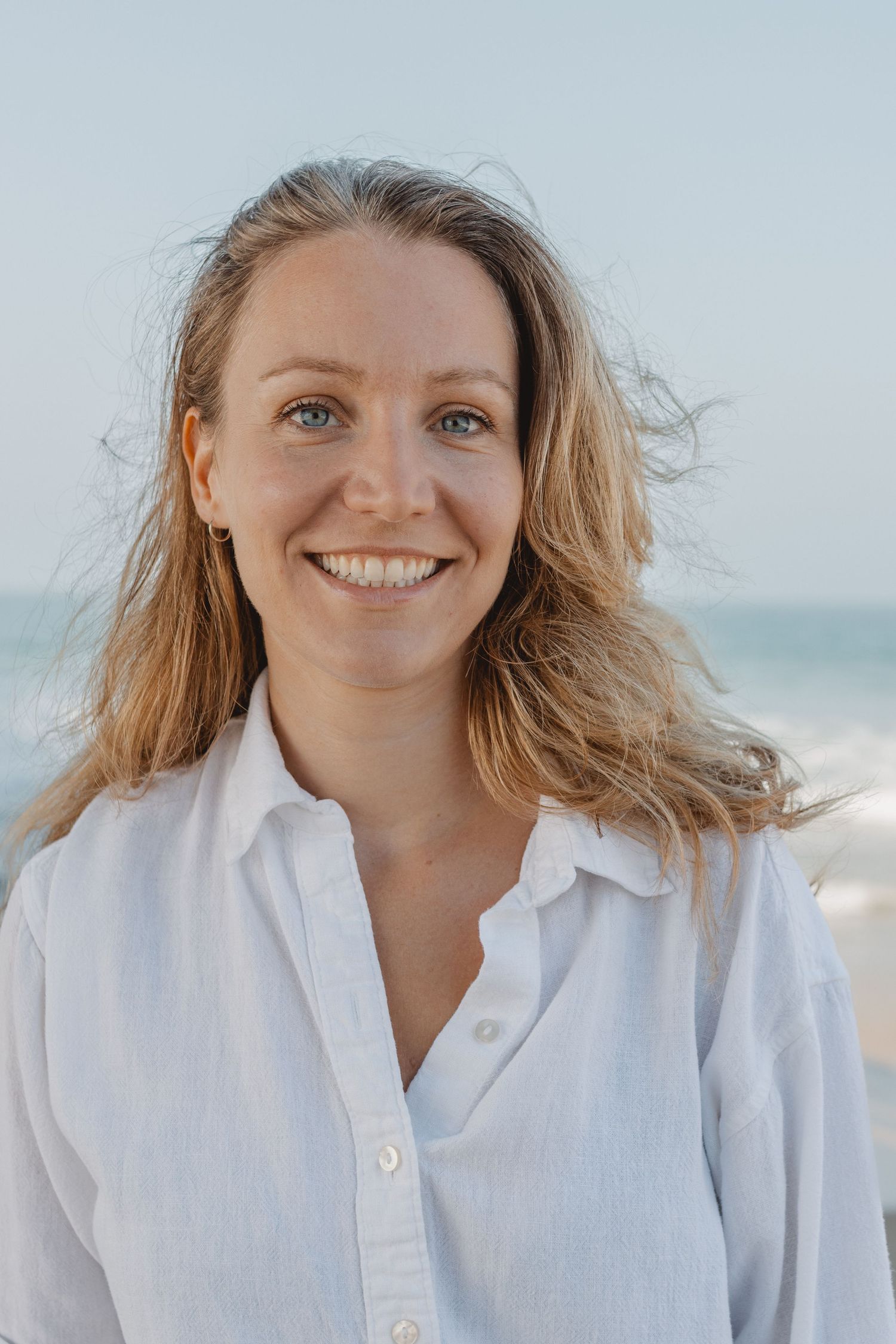 A person in a white button-down shirt smiles warmly against a beachy background with soft natural lighting in Newport Beach at the Pier.