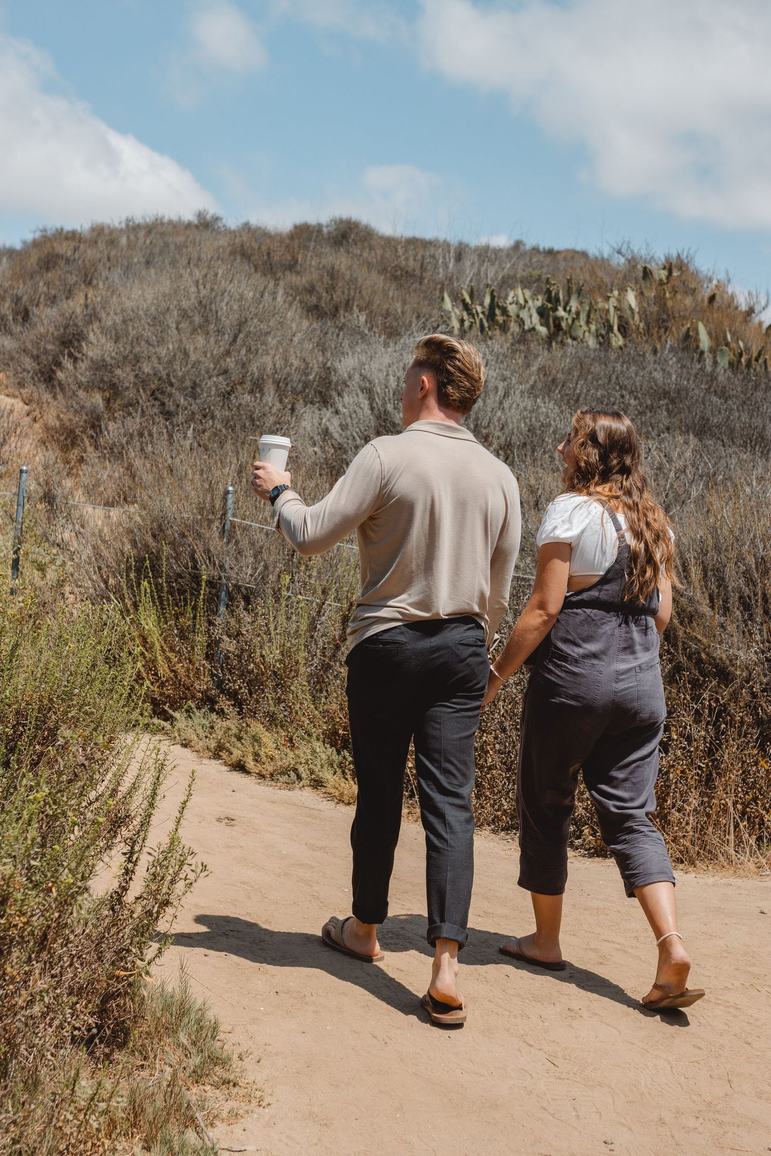 A pregnant couple walk along a dirt path on a hillside in Newport Beach enjoying a scenic overlook on a sunny day.
