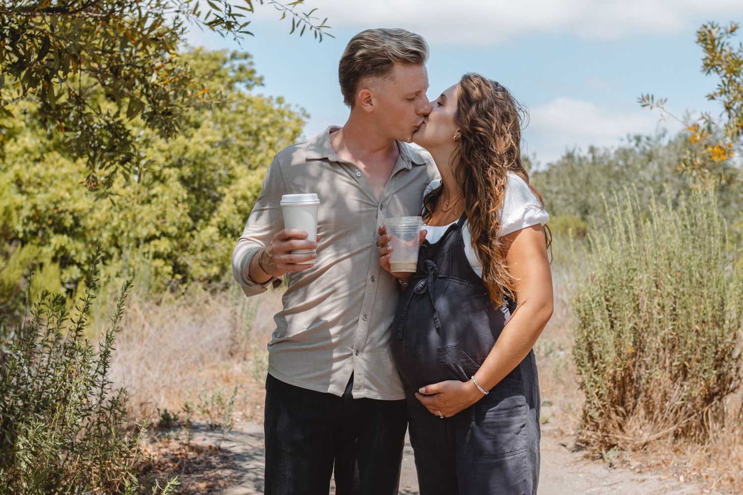 A pregnant couple stands together in a sunlit field of tall grass and wildflowers in Orange County sharing a romantic moment together.